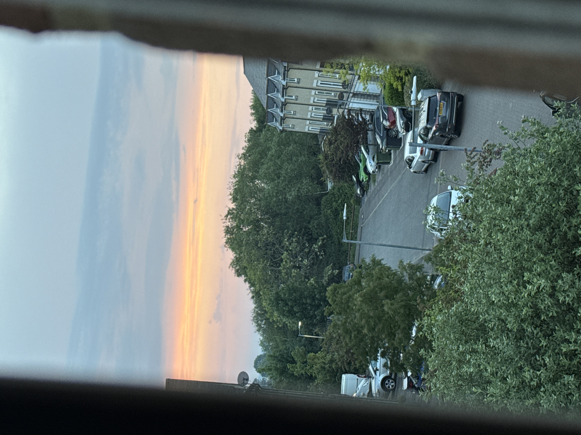 From a high vantage point within a building in Great Yarmouth, United Kingdom, a view extends towards a streetscape. The sky, transitioning from light blue to golden hues, suggests either dawn or dusk. A row of terraced houses stands adjacent to the street, with parked cars lining the road. Trees densely populate the area between the buildings and the foreground. Street lamps are visible, as are satellite dishes affixed to buildings. The vantage point is partially obscured by a structural element, such as a window frame.