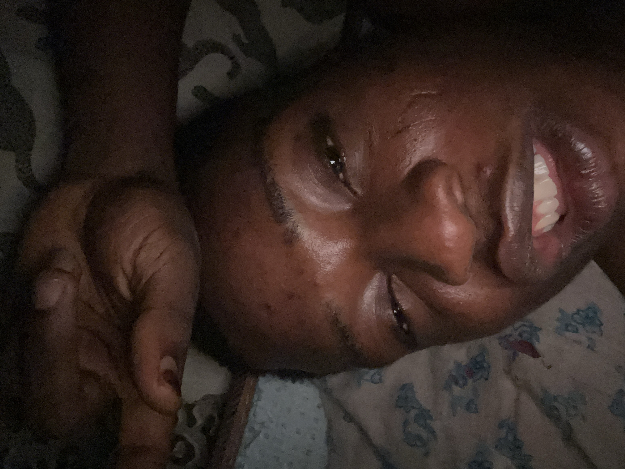 A close-up, low-angle shot shows a dark-skinned person lying on what appears to be a patterned bedsheet. The person's face is turned towards the camera with a partial smile revealing their teeth. One hand is visible in the upper left corner of the frame. The lighting is dim, casting shadows across the scene. Location: Unknown, Unknown.