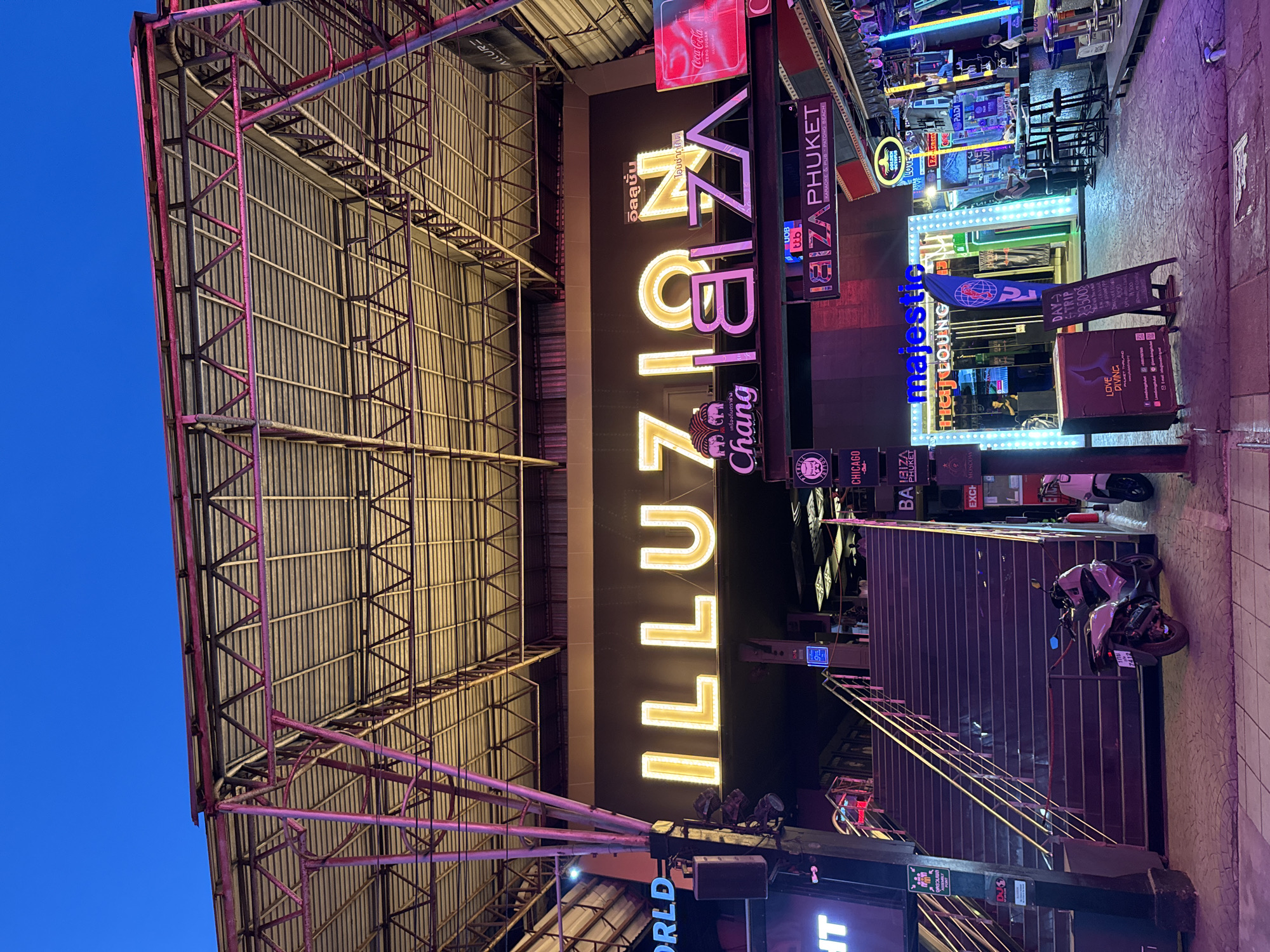 The image depicts a nighttime scene on a street in Patong, Thailand, with a focus on the entrance to a club or entertainment venue. The dominant structure is a large, open-air building with a prominent illuminated sign that reads "ILLUZION IBIZA" in large, yellow letters. Below this, smaller neon signs display "Chang" and what appears to be Thai script. The building's facade is dark, contrasted by the bright signage and colorful neon lights.
A wide set of dark-colored stairs leads up to the entrance, flanked by metal railings. A few people are visible on the stairs and at the entrance, appearing to be patrons or staff. To the left of the stairs, a motorcycle is parked on the sidewalk, and to the right, another scooter is partially visible.
The surrounding environment shows typical street elements of a lively nightlife district. To the right of the main entrance, there are more illuminated signs for businesses like "Majestic Raj Lounge," "Ibiza Phuket Karaoke & Pub," and "Golden Sands." A bar area with stools is set up outdoors, with several people standing or sitting at the bar. The street is paved with patterned tiles, and the overall lighting suggests a vibrant, albeit somewhat gritty, urban setting. The sky above is a deep twilight blue. The metal framework of the building's roof, with its exposed beams and corrugated roofing, is visible overhead, illuminated with pink and purple lights.