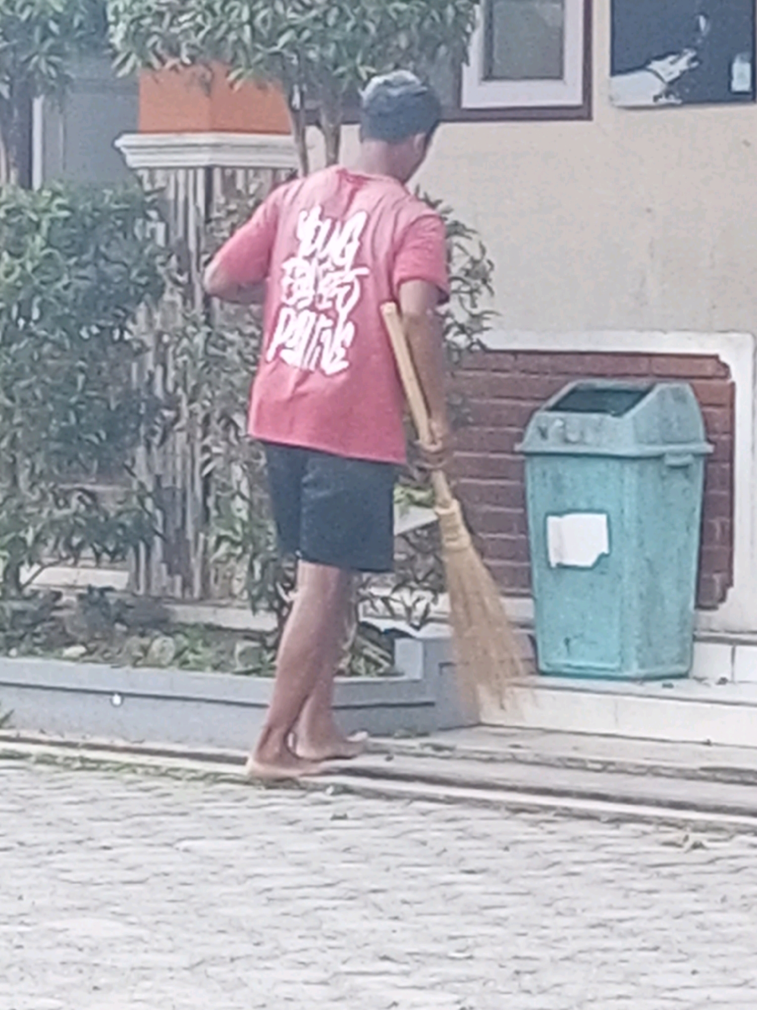 In the village of Krasak, Indonesia, a person is seen from behind, wearing a pink t-shirt with white text and dark shorts. They are barefoot and are sweeping the ground with a broom. To their right, there is a turquoise trash can with a white label. The background shows a building with windows and a brick wall, surrounded by greenery.