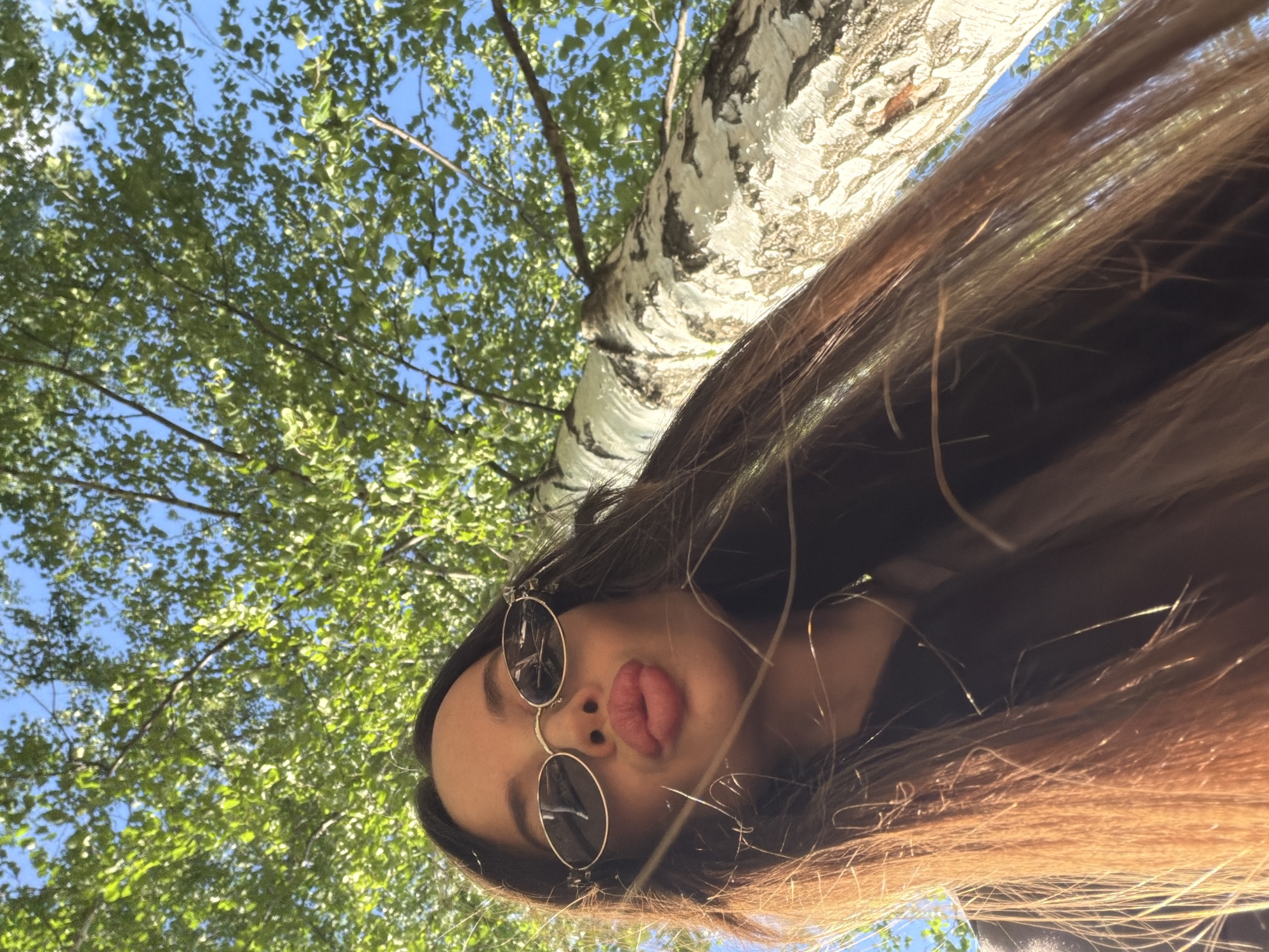 A low-angle, upward-facing shot captures a female subject posing playfully outdoors, likely in Izhevsk, Russia. The scene is dominated by a clear blue sky and a lush canopy of green leaves from a tree, with a distinctive white and black-patterned birch tree trunk visible on the left side of the frame.
The main subject is a young woman, seen from the chin up, with her head tilted to the right. She has long, flowing brown hair that cascades across the lower and right portions of the image, appearing slightly wind-blown or artfully arranged. She is wearing round-rimmed sunglasses, reflecting the bright sky, and is making a "kiss" face with her lips pursed outwards. The lighting suggests a bright, sunny daytime, casting highlights on her hair and the tree bark. There is no visible text in the image.