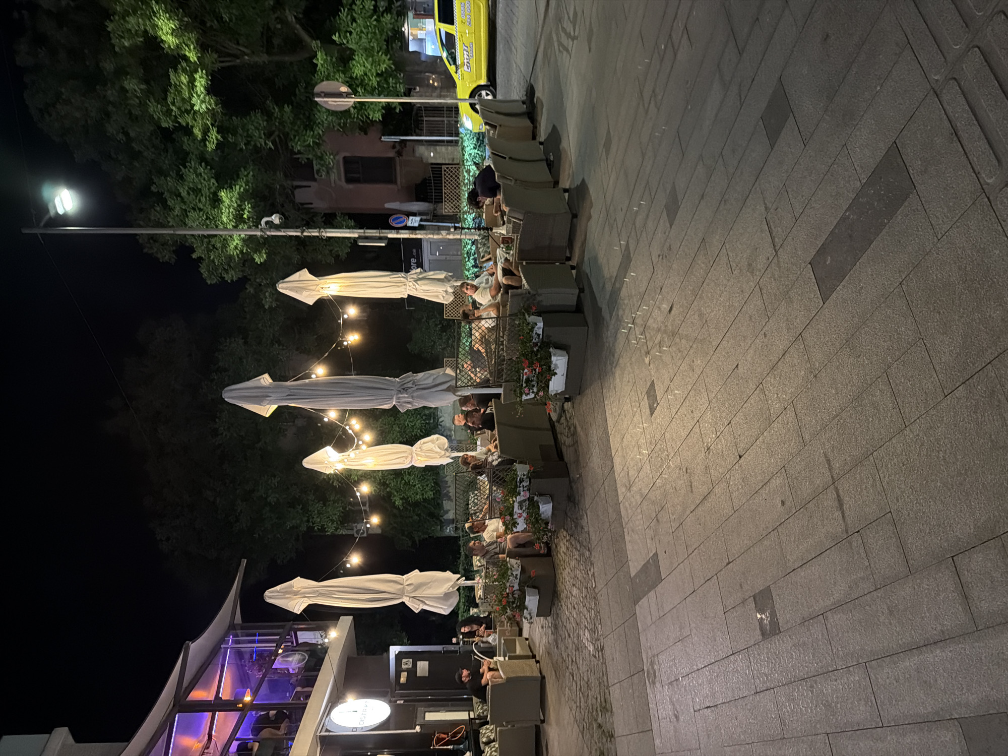 This image depicts an outdoor seating area of a establishment, likely a restaurant or cafe, at night in Burgas, Bulgaria. The scene is illuminated by string lights and a street lamp, casting a warm glow on the pavement and surrounding trees. Several people are seated at tables and on comfortable lounge chairs, engaged in conversation or relaxation. Large, closed umbrellas are positioned around the seating area, suggesting they provide shade or protection from elements. In the background, a yellow taxi cab is visible on the street, indicating an urban setting. The architecture of the building suggests a modern design with glass elements and subtle interior lighting. The overall atmosphere appears to be one of a pleasant evening social gathering.