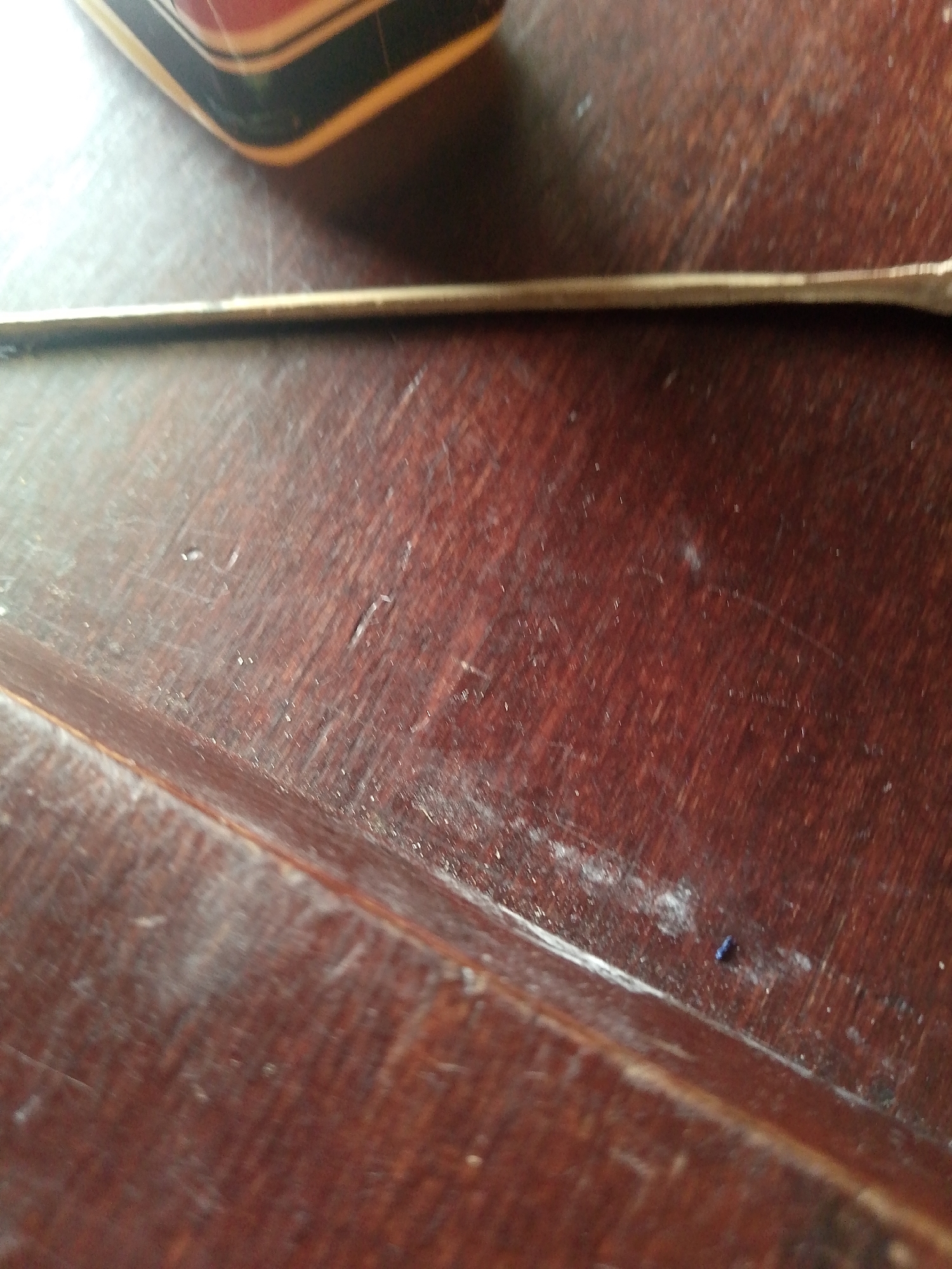 The image shows a close-up view of a wooden surface, possibly a table or desk, with a thin, metallic object, likely a ruler or a similar flat, elongated item, placed horizontally across it. The wooden surface exhibits a rich, dark brown color with visible grain patterns and numerous scratches, indicating wear and age. A portion of another object, with curved edges and distinct layers of color (black, red, and yellow-ish gold), is visible in the upper left corner, out of focus. In the lower right quadrant, a small, dark blue speck, possibly a piece of debris or a mark, is present on the wooden surface. The lighting in the scene appears to be overhead, casting subtle shadows and highlighting the texture of the wood. There is no visible indication of Jauro Yinu, Nigeria in the image.