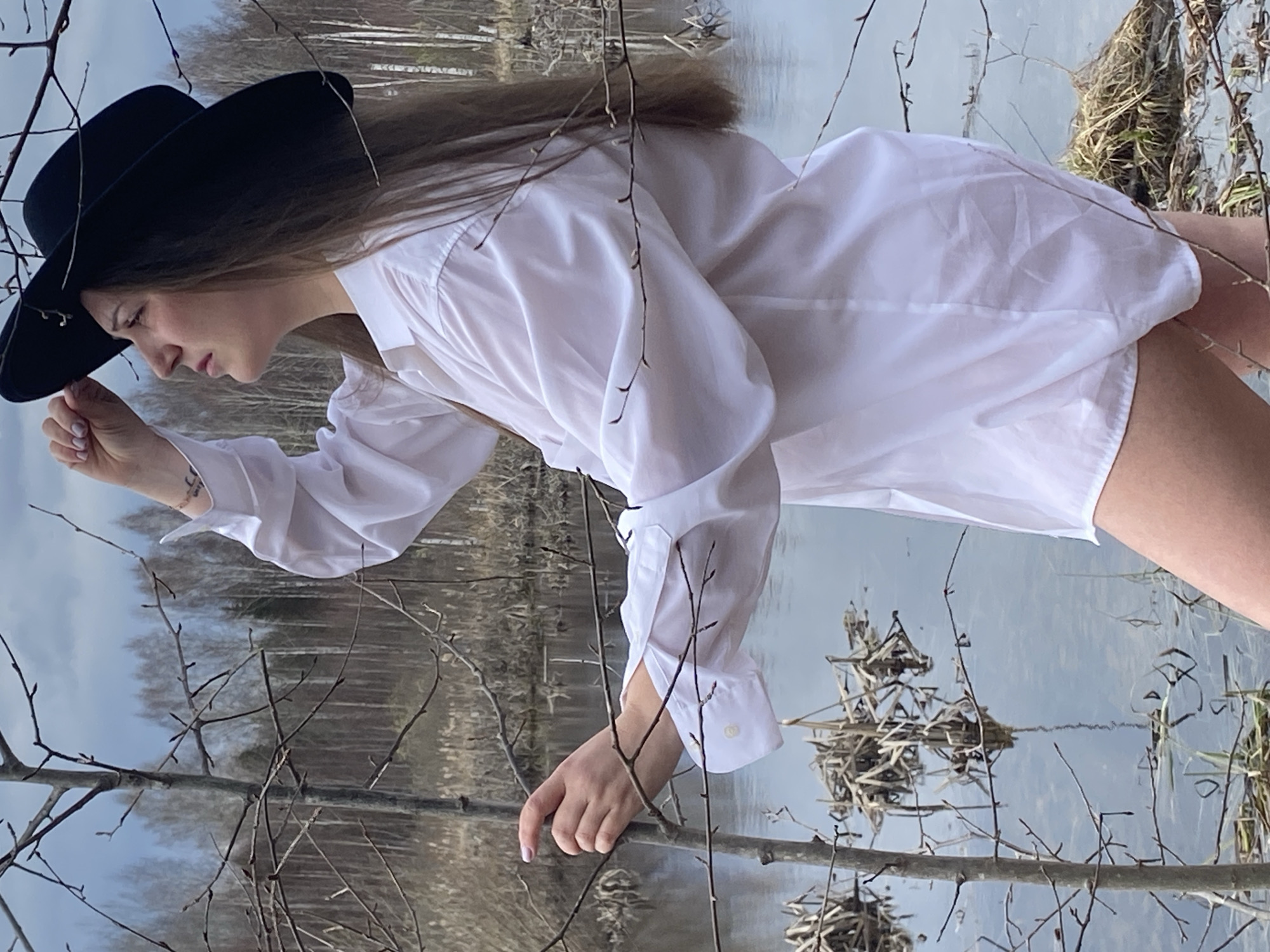 A woman stands near a body of water in what appears to be a natural environment. She is wearing a white, long-sleeved shirt and a black hat. Her body is angled diagonally in the frame, with her left hand reaching toward a tree branch. The background includes trees, water, and what looks like natural debris in the water. The scene may be in Марковский, Russia, based on the prompt.