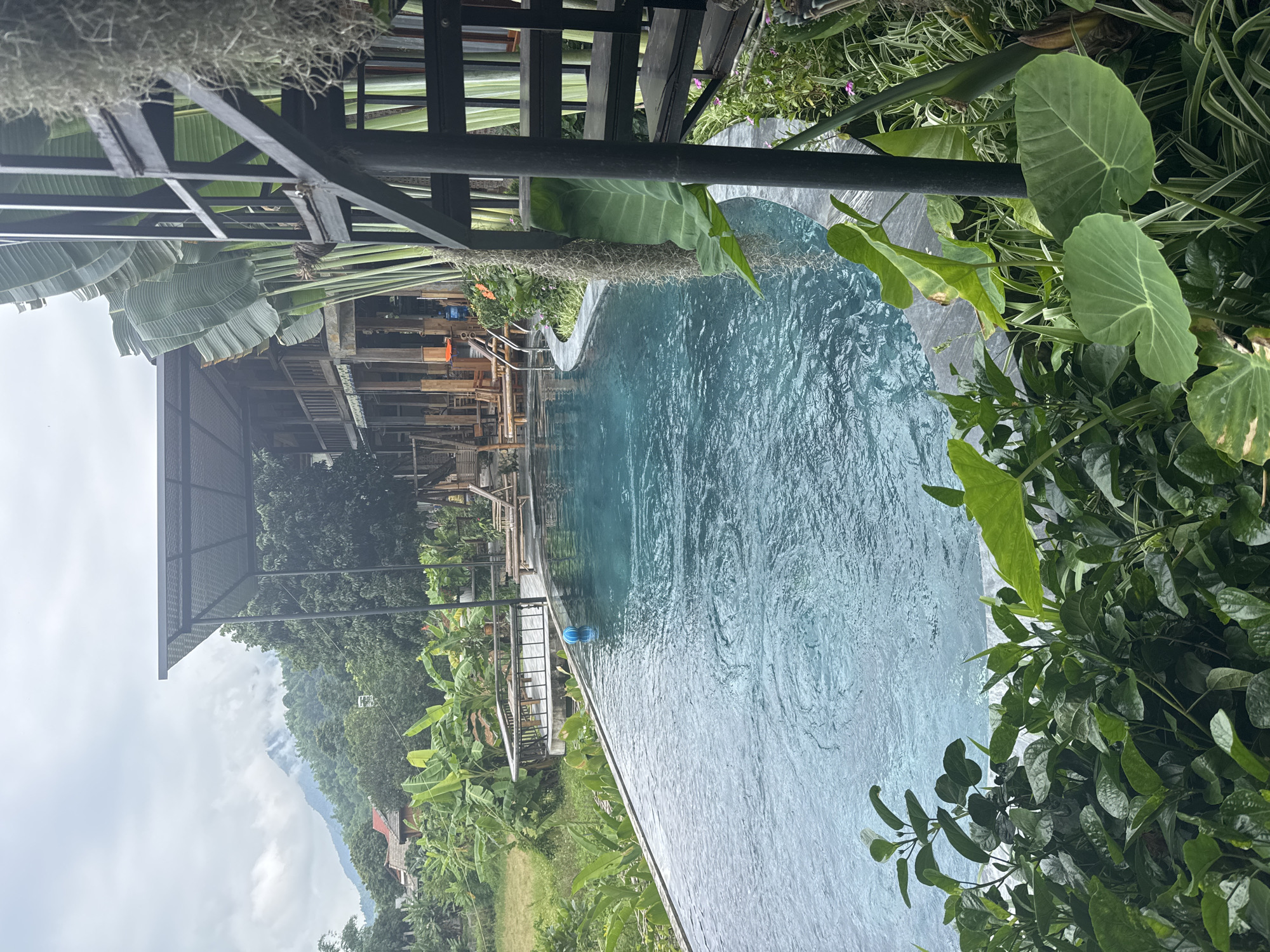 An infinity pool with turquoise water is the central feature of this image, situated outdoors in a tropical environment. The pool appears to be part of a resort or hotel in Mai Chau, Vietnam, given the lush vegetation and mountainous backdrop visible in the distance. A spiral staircase with black metal railings leads up to a multi-story wooden building with an open-air design, suggesting a blend of traditional and modern architecture. Large, tropical plants, including banana trees and elephant ear plants, surround the pool and building, enhancing the serene and natural ambiance. The sky is overcast, indicating a potentially humid or rainy day, common in the region. There are no people visible in the image, making it a tranquil scene focused on the interplay between the man-made structure and the surrounding natural landscape. The water in the pool has visible ripples, suggesting slight movement, and a blue ball floats near the edge. The overall impression is one of a peaceful, secluded retreat immersed in nature.