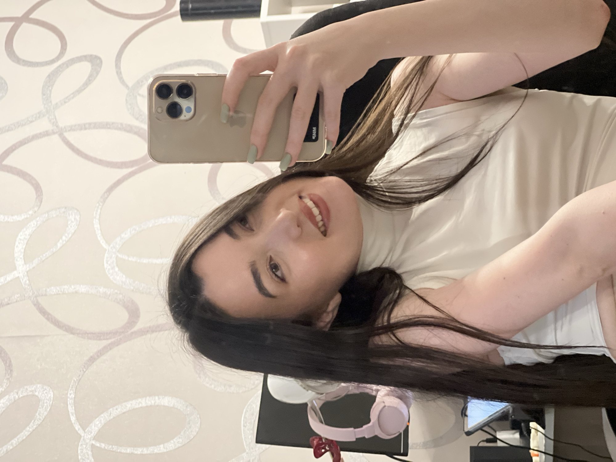 The image is a selfie taken indoors by a young woman with long dark hair and light skin. She is wearing a sleeveless off-white top. Her right hand holds a gold-colored smartphone, with which she is taking the picture. Her nails are painted a light green color. In the background, there is a wall with a light-colored wallpaper featuring a swirling pattern in pink and silver. To the left of the woman, a computer monitor is visible, with pink headphones hanging on it. To the right, a white shelf unit and part of a black chair are present. The lighting suggests an indoor environment, likely a room in a home or office. There is no indication of the location being in the city of Kazan, Russia.