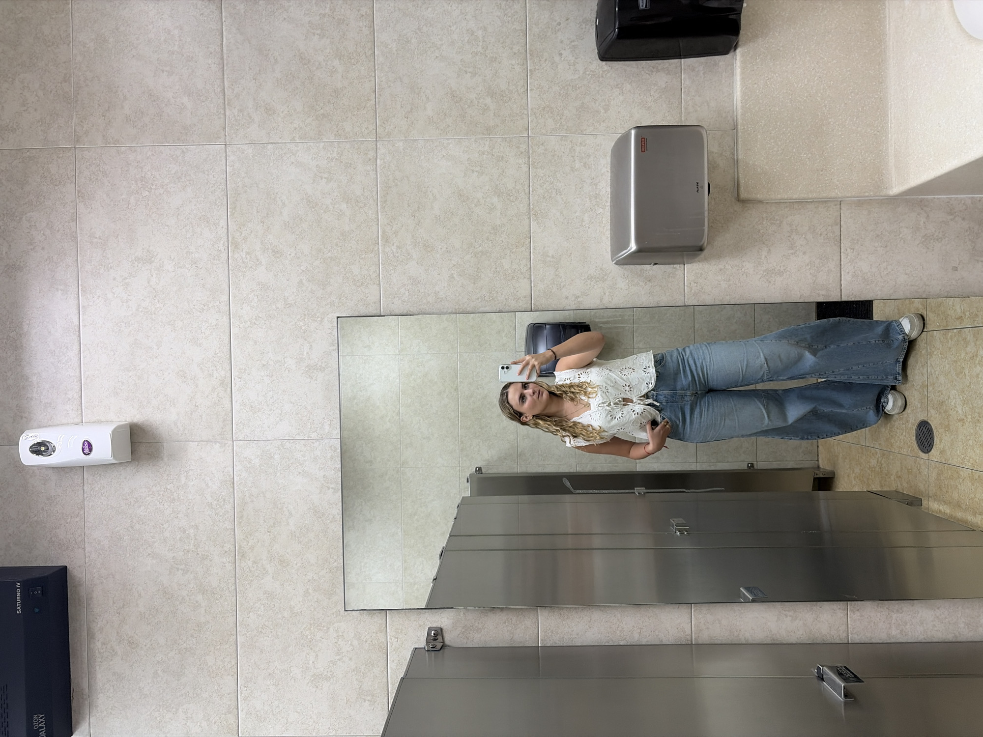 The image shows a woman taking a selfie in a public restroom. The restroom has light beige tiled walls and floors. The woman is standing in front of a mirror, holding a smartphone. She is wearing a white blouse and blue jeans. The restroom features several amenities including a paper towel dispenser, a hand dryer, and multiple stainless steel stalls. The city of San Andrés Cholula, Mexico, cannot be determined from the information in the image.