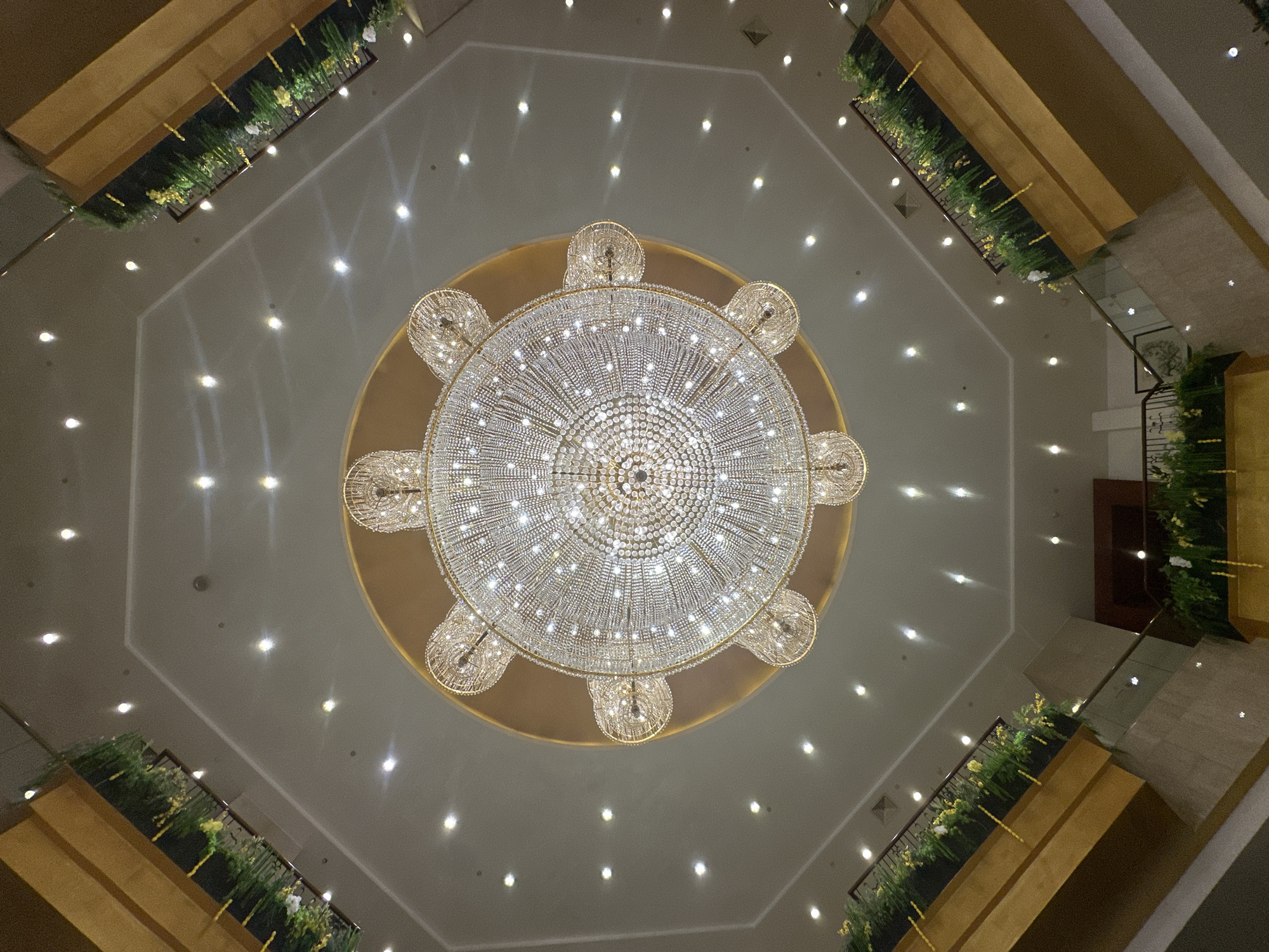 This is a low-angle, wide shot of an indoor atrium featuring a large, ornate crystal chandelier as the central focus. The chandelier is suspended from a gold-colored circular ceiling medallion and is adorned with numerous sparkling crystals and lights. The ceiling itself is octagonal and recessed, with a lighter colored surface. Numerous small, bright lights are embedded in the ceiling, creating a star-like effect.
Surrounding the central area are multi-level walkways or balconies, constructed with polished wood and stone. These walkways are lined with railings and feature planters filled with lush green foliage and yellow and white flowers. The architecture suggests a grand interior space, possibly a hotel lobby, convention center, or a large commercial building.
The image does not contain any people or specific identifiable landmarks that would indicate a precise location within Kunming, China. The scene is one of interior architectural design and lighting.