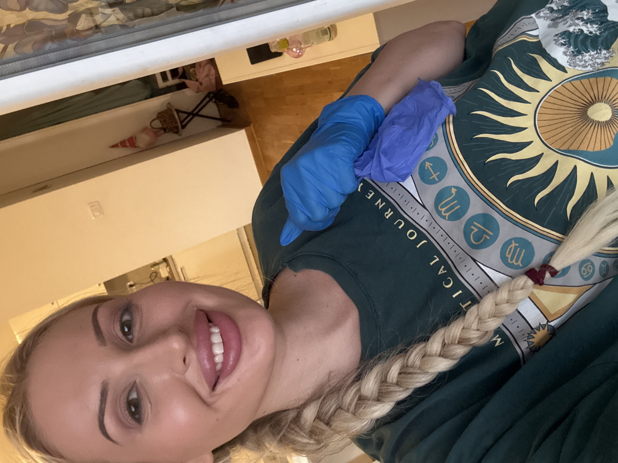 A blonde woman with a braid is taking a selfie in an indoor environment. She is wearing a dark green t-shirt with a celestial design featuring a sun and zodiac symbols. She is also wearing blue nitrile gloves and holding another pair of purple nitrile gloves in her right hand. The background shows a room with light-colored walls, a wooden floor, and some furniture, including a white cabinet with items on top and a small table with a decorative gnome. The lighting is warm and artificial.