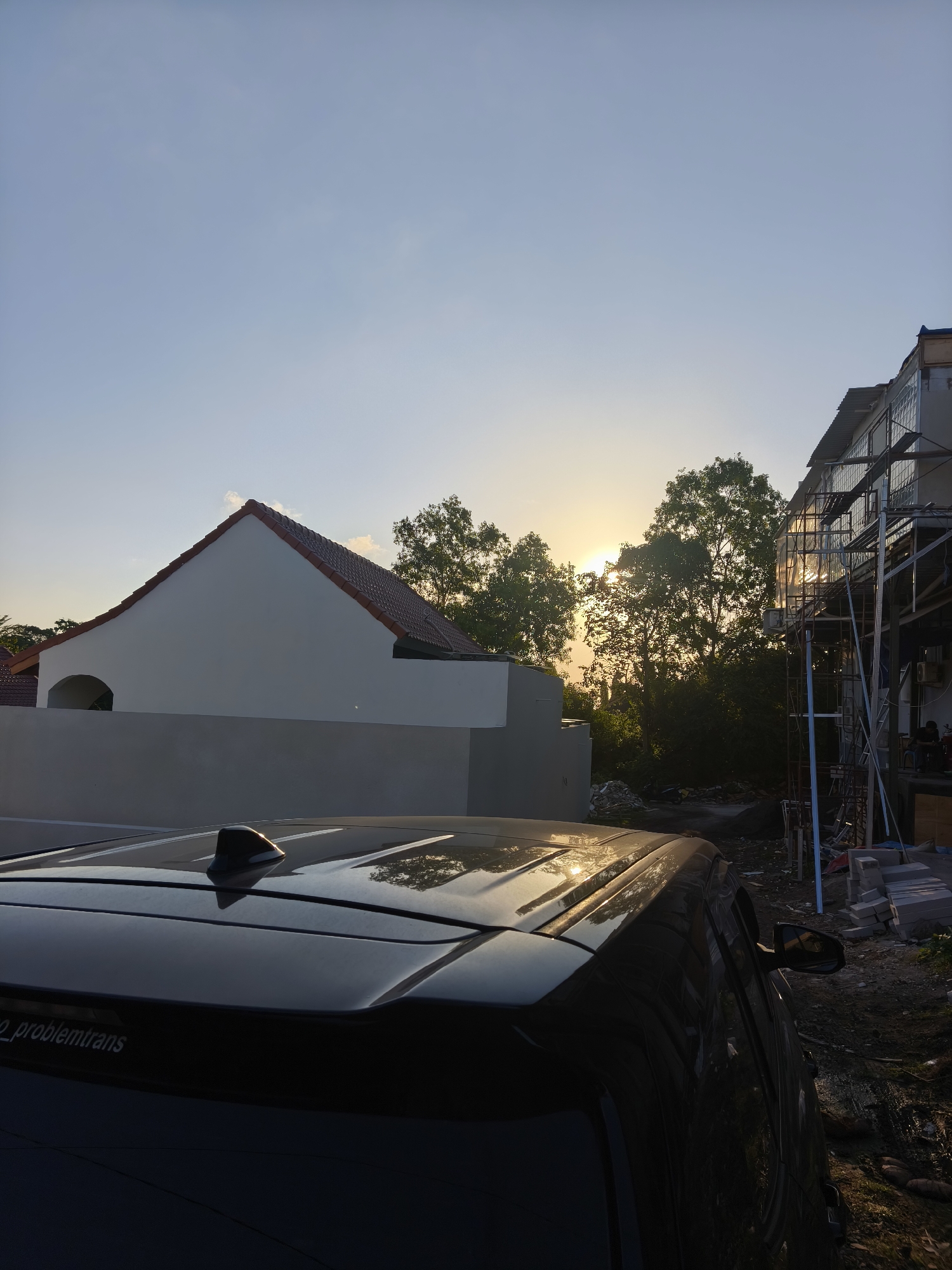 The image captures a sunset scene in Kuta Selatan, Indonesia. In the foreground, the dark, glossy roof of a car is visible, reflecting the warm glow of the setting sun. A shark fin antenna is mounted on the car's roof. In the midground, a light-colored building with a tiled, sloped roof and an arched entryway stands to the left. To the right, a building under construction is partially visible, with scaffolding and construction materials. Dense foliage and trees are situated between the buildings, with the sun peeking through the branches. The sky is a gradient of light blue to pale yellow near the horizon, indicating the late afternoon or early evening. The ground appears to be unpaved, with some construction debris and dirt. The overall atmosphere is tranquil, with the natural light creating a warm and somewhat dramatic effect.