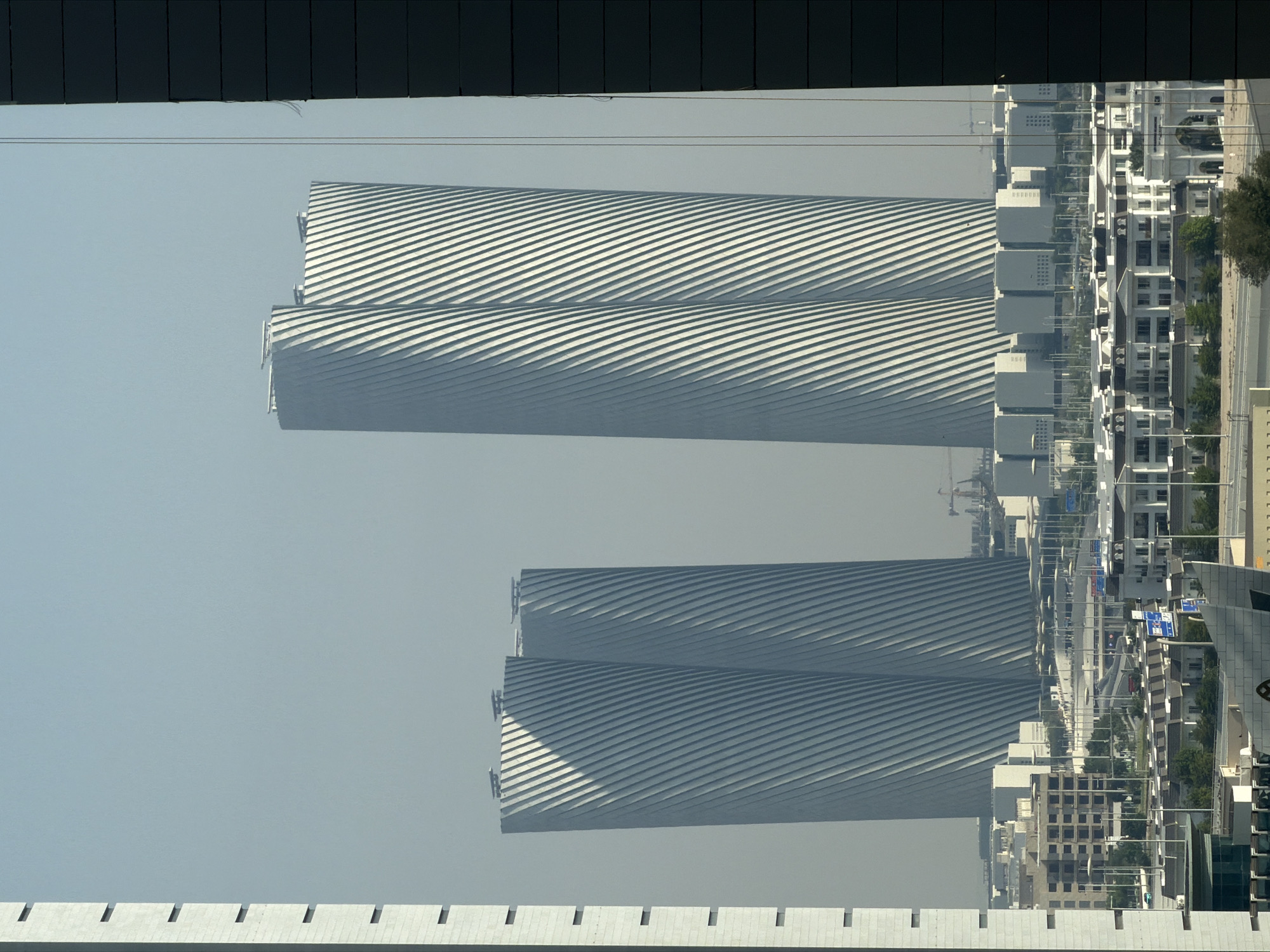 The image depicts a cityscape with a focus on modern architecture. In the background, two distinctive, tall, cylindrical skyscrapers with a twisted, ribbed facade dominate the view. To the left of these, a closer, similarly shaped skyscraper is partially visible, also exhibiting the ribbed design. In the foreground and mid-ground, a dense urban landscape unfolds, characterized by numerous low-rise buildings with white facades, arranged in a grid-like pattern. These buildings appear to be residential or commercial complexes, with visible windows and architectural details. Streets and roads are visible, with some indication of vehicular traffic, though not clearly discernible. The environment is characterized by a hazy, light blue sky, suggesting a clear but perhaps dusty or humid atmosphere. The overall impression is one of a contemporary, developed urban environment.