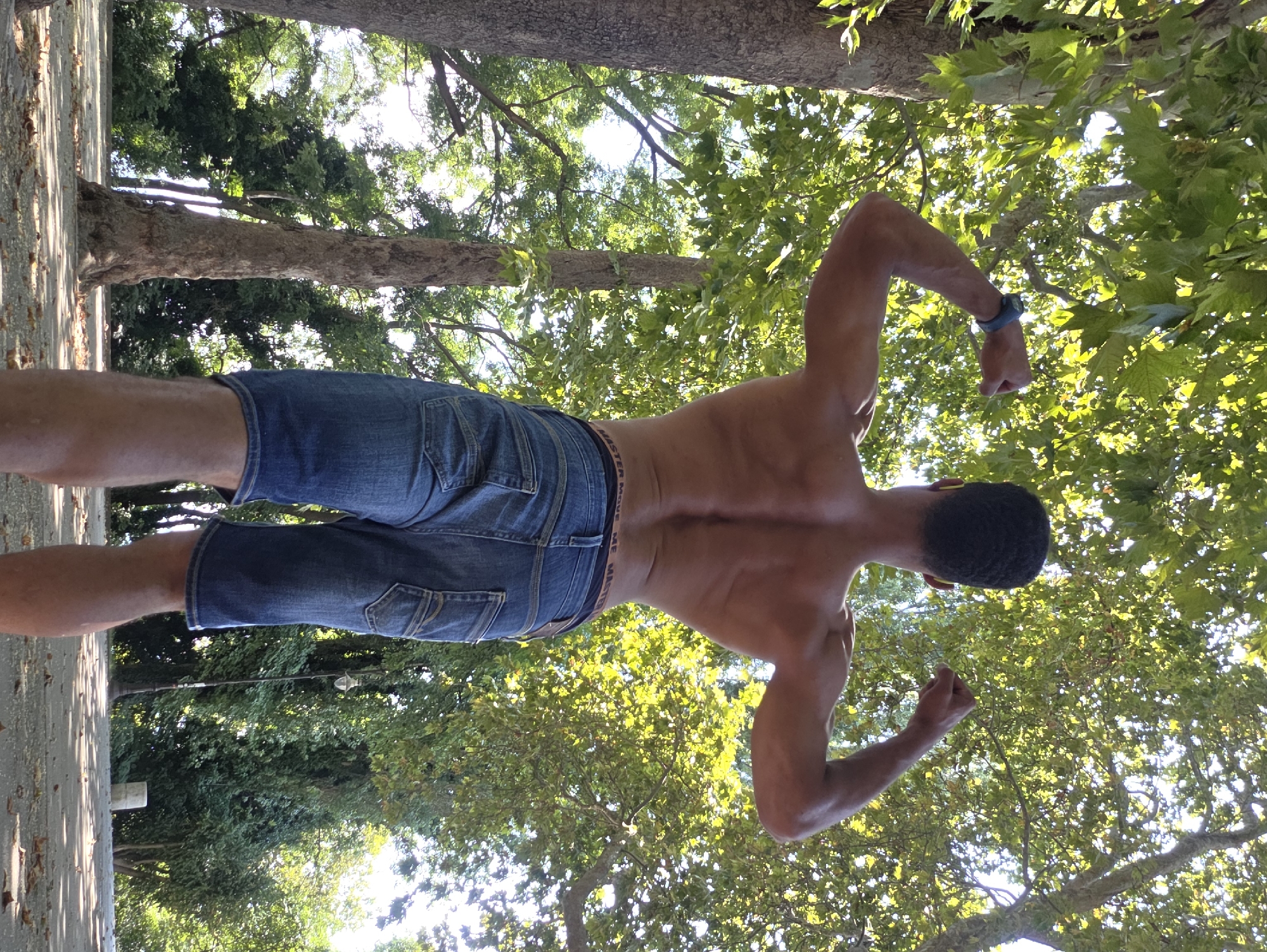 A man, viewed from behind, stands outdoors, flexing his biceps. He is wearing denim shorts and a watch on his left wrist. The setting appears to be a park or garden with trees and foliage. The lighting suggests it is daytime. The background includes trees, leaves, and a pathway. The image quality is clear. No specific landmarks of Varna, Bulgaria are discernible.