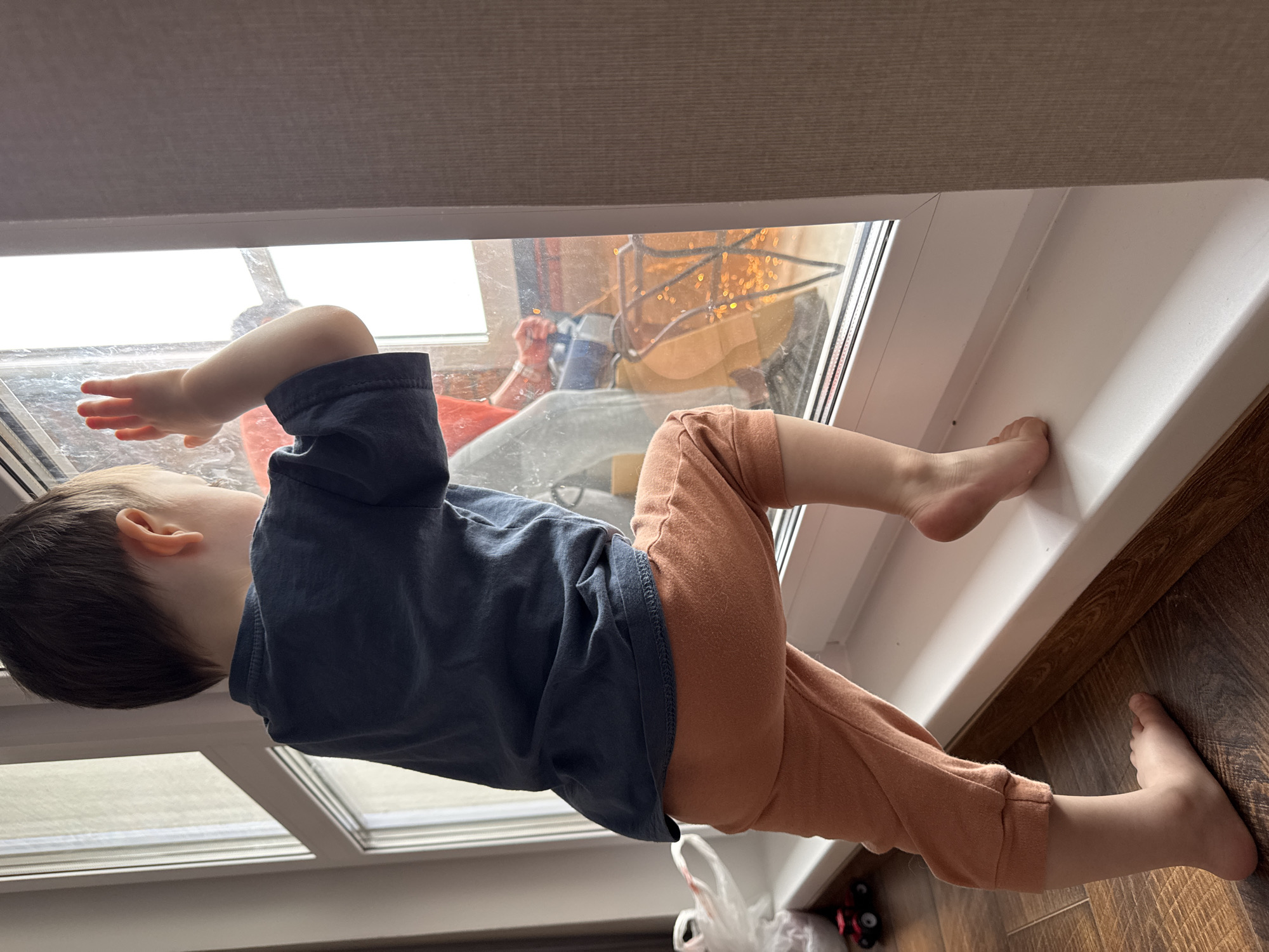 A young child is standing barefoot on a wooden floor next to a white window frame. The child wears a navy shirt and salmon-colored pants. The child is facing the window and appears to be looking outside with one hand pressed against the glass. Outside the window, there is a blurry view of what appears to be a person sitting in a chair with a yellow object above them. The window is partly obscured by a window shade above. A plastic bag and a small toy are on the floor near the child's feet. The location is not explicitly stated as Izhevsk, Russia and cannot be determined from the image.