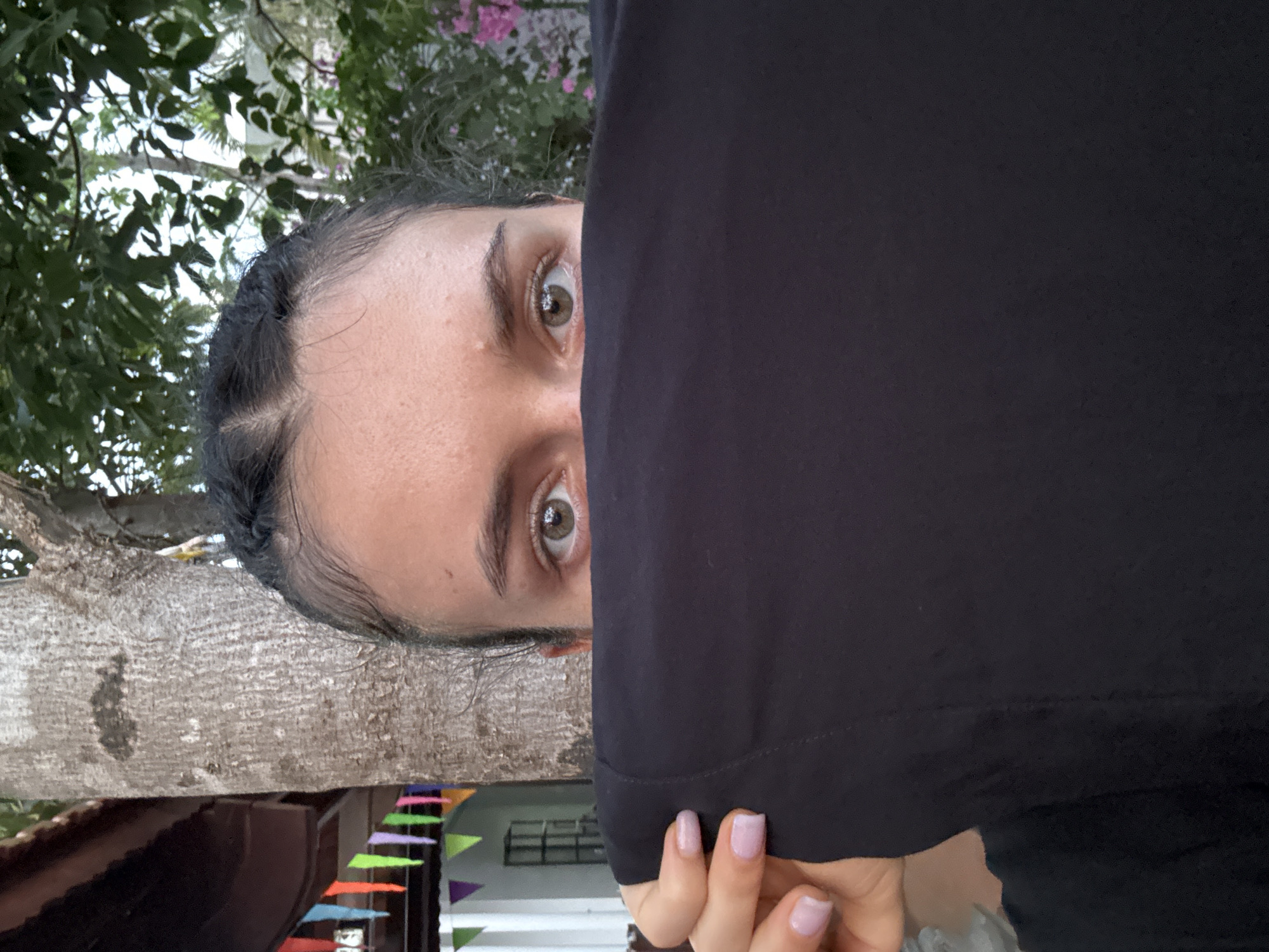 A close-up photograph, rotated 90 degrees counter-clockwise, focuses on a person’s face, partially obscured by a black fabric, possibly a shirt or garment. The face, with light skin and fair eyes, is visible from just above the eyebrows to the nose. Braided hair recedes from the forehead. The right side of the face is mostly hidden behind the black fabric, with a manicured hand visible in the lower-right corner. The background includes a tree trunk and foliage, along with decorative colorful triangular flags in the mid-ground, suggesting a festive atmosphere. Based on visual elements, the location could be the city of Mũi Né, Vietnam, given the prevalence of palm trees and the style of decorations.