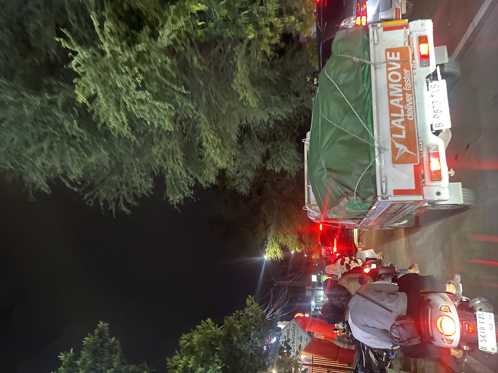 At night in North Jakarta, Indonesia, a Lalamove delivery truck laden with a green-covered load is parked in traffic. Behind it, motorcyclists are waiting. Streetlights and storefronts are visible. A sign for "Samudra barbershop" is also seen. The motorcycle closest to the camera has a license plate that reads B 5418 FXO. The truck's license plate reads B 8873 TIS.