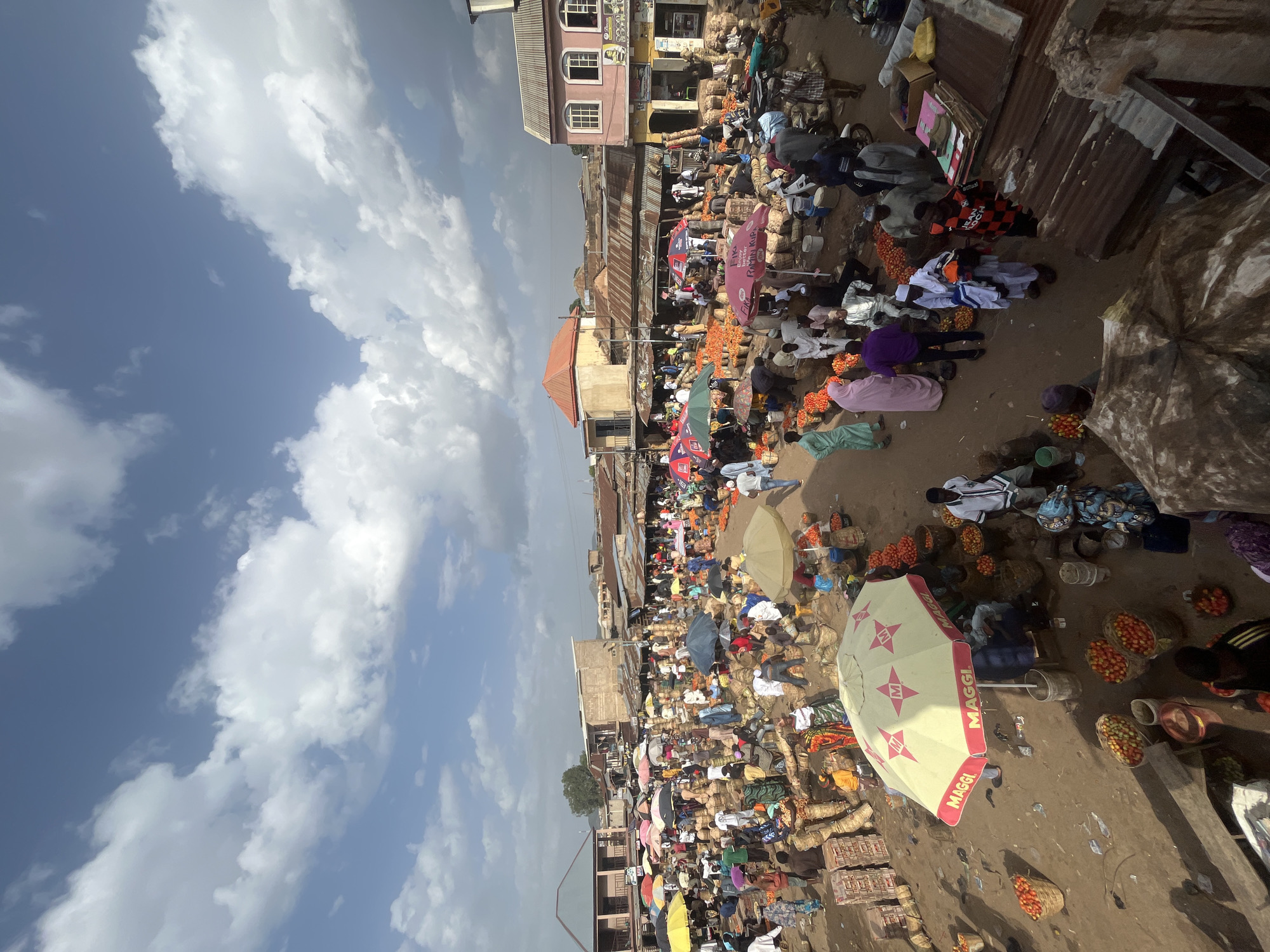 The image depicts a busy outdoor market in Rafin Par, Nigeria. The market is filled with people, many of whom are vendors or shoppers. The ground is dusty and uneven, with baskets and sacks of produce, primarily tomatoes, scattered throughout. Several stalls are set up, with vendors under umbrellas or awnings. In the background, there are various buildings, some with corrugated metal roofs, indicating a mix of permanent and temporary structures. The sky is partly cloudy, with a mix of blue sky and white clouds. The overall scene suggests a vibrant and active marketplace, typical of many rural or semi-urban areas in Nigeria.