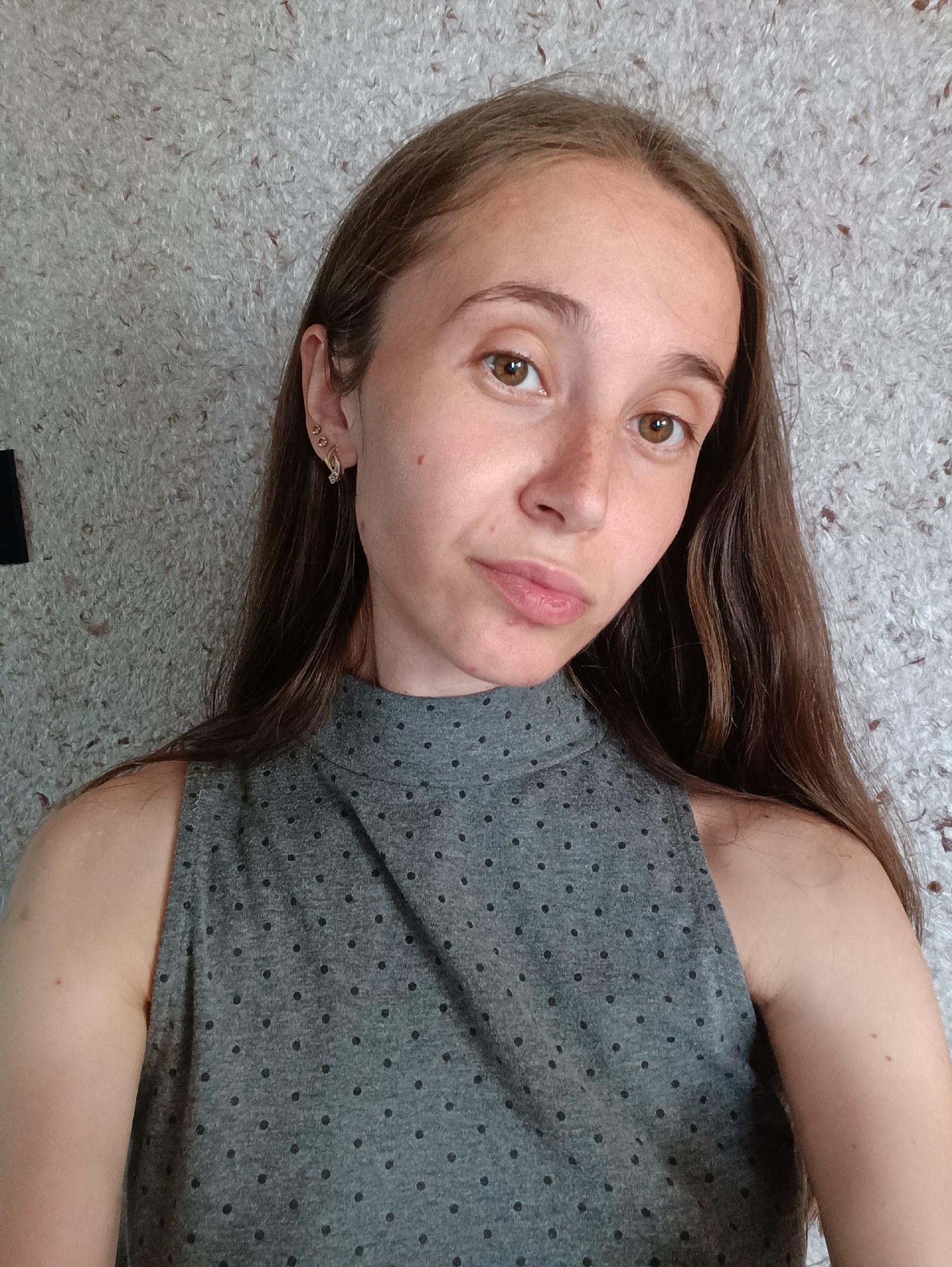 A young woman with long, brown hair and light brown eyes is pictured in a close-up, slightly angled selfie. She is wearing a grey sleeveless top with a high neckline and a subtle dotted pattern. Her left ear is adorned with two small earrings: one a cluster of three small, light-colored stones, and the other a more elaborate gold hoop with a decorative element. The background is a textured, light-colored wall. The image appears to have been taken indoors.