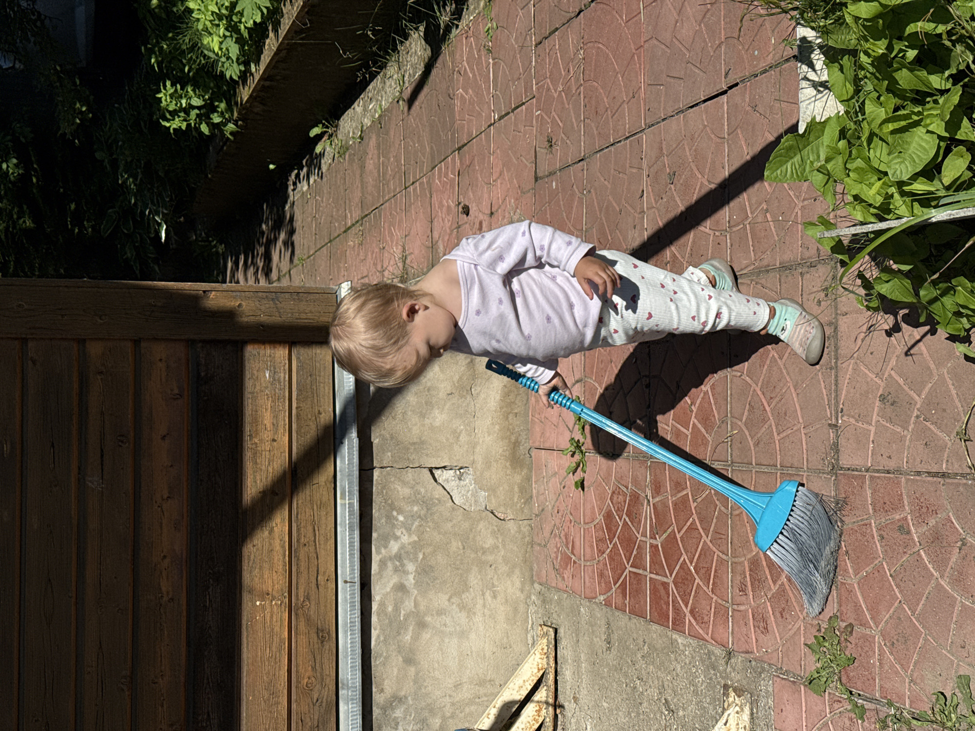 A young child with blonde hair, wearing a light purple long-sleeved shirt with a floral pattern and white pants with red hearts, stands on a red tiled pathway next to a weathered wooden building in Дзержинец-83 Снт, Russia. The child is holding a bright blue broom with gray bristles, appearing to be sweeping. Sunlight casts a shadow from the building onto the pathway and the child. To the right of the pathway, there is a low, dark planter box filled with lush green plants and foliage. The ground is cracked in some areas, and there is debris on the pathway.
