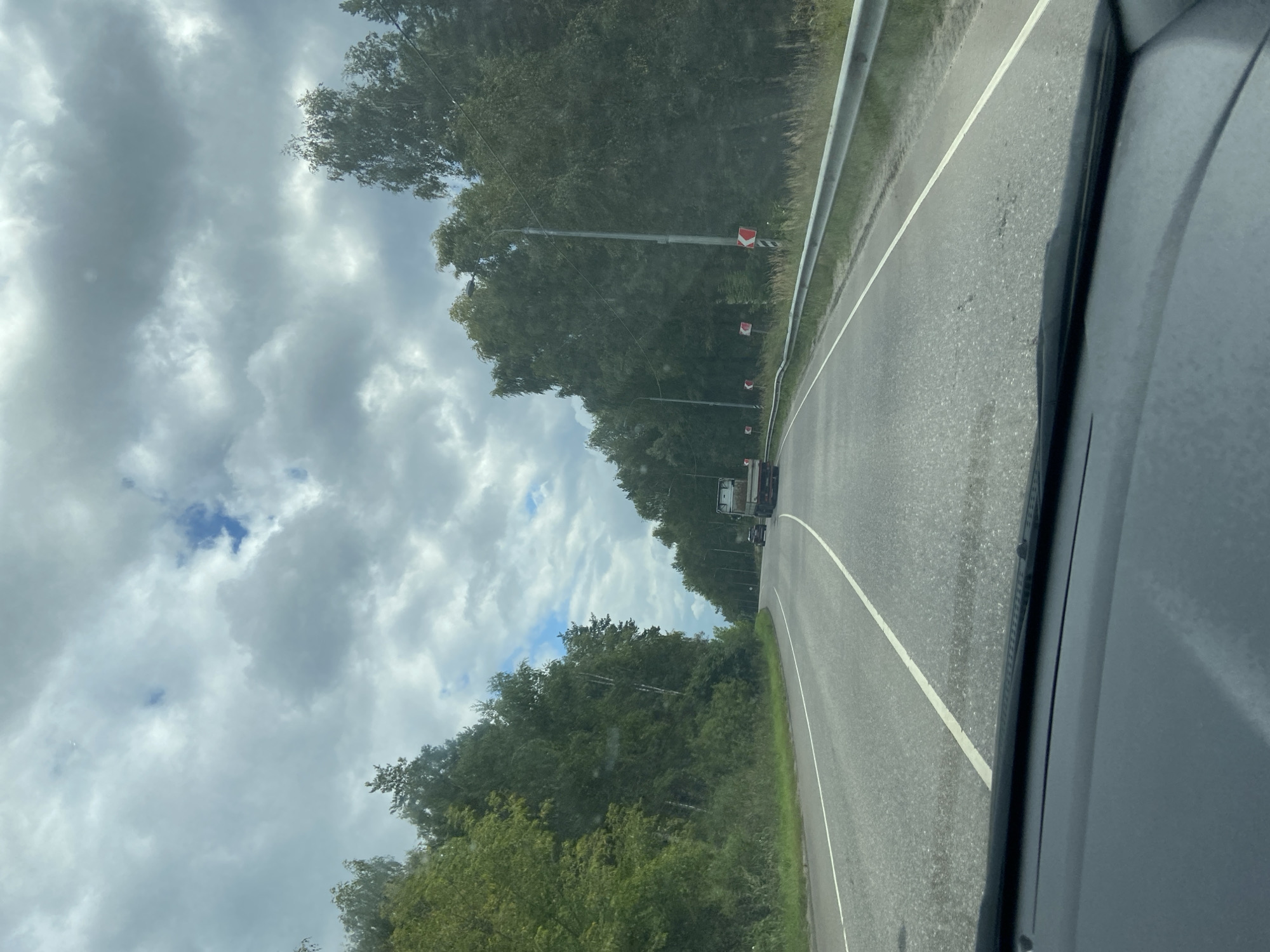 The image captures a view from a vehicle traveling on a two-lane asphalt road bordered by dense green trees. The sky is overcast with scattered clouds, allowing some patches of blue to be visible. The road curves to the right. Traffic signs indicating a curve ahead are visible along the right side of the road. A truck and a car are present on the road, moving in the same direction as the viewer. The foreground shows the dashboard and windshield of the vehicle, implying the photograph was taken from the driver's or passenger's seat. The location is the city of Alekseevskaya, Russia.