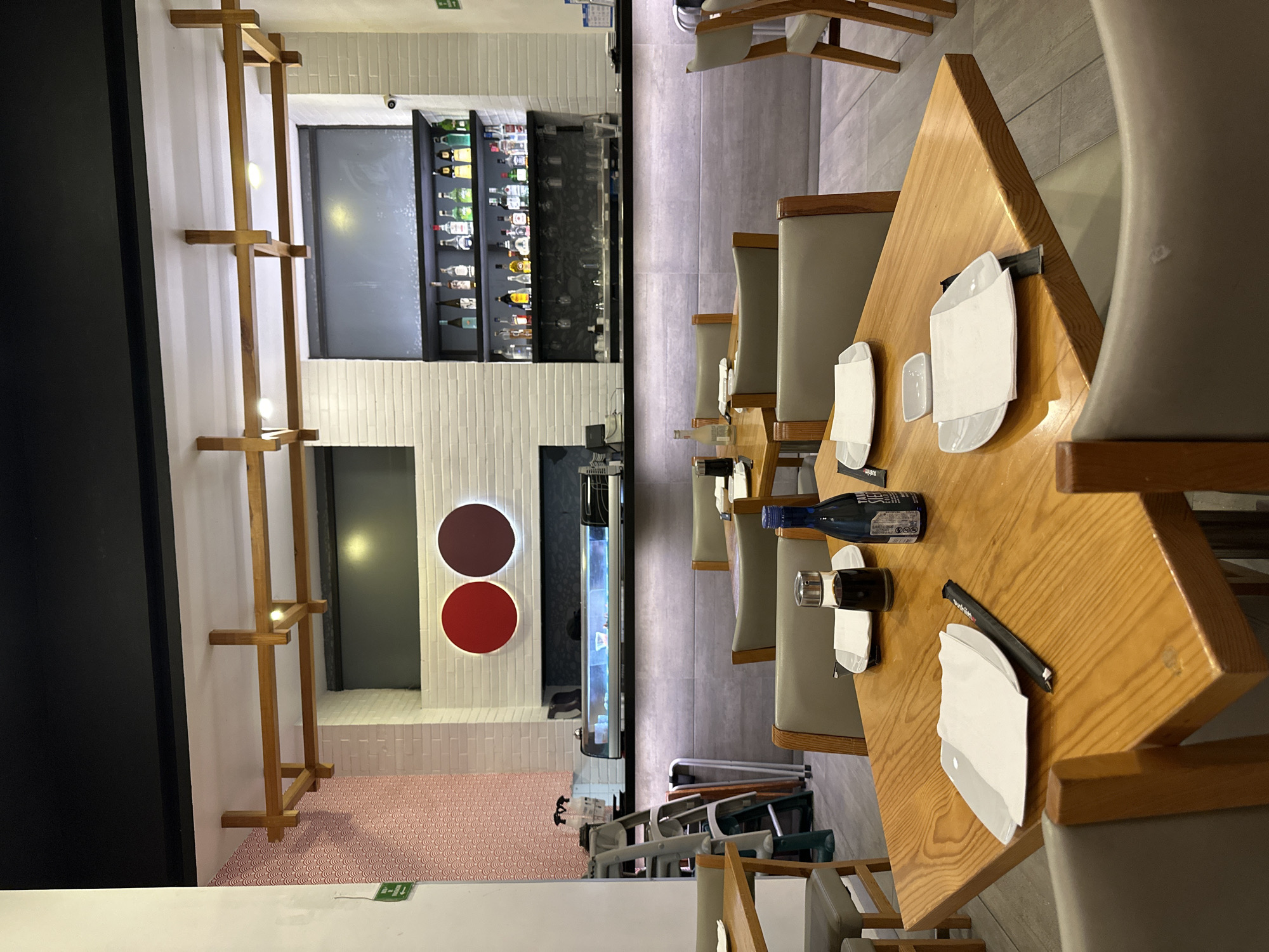 This image captures the interior of a contemporary restaurant or bar, likely located in Mexico City, Mexico. The scene is presented from a slightly elevated perspective, showcasing a dining area and a bar section.
In the foreground and extending towards the center-right, several square wooden tables are arranged with light grey upholstered chairs. Each table is meticulously set with white rectangular plates, folded white paper napkins, and black chopstick wrappers (some with "Sushi" visible). Condiment dispensers and two bottles – one blue with "PREMIUM" text and another dark brown – are also placed on the tables.
To the left, a long, well-stocked bar counter features a dark surface. Behind it, backlit shelves display a wide array of liquor bottles, suggesting a full-service bar. Below the bar, a narrow, illuminated display case or aquarium is visible. The walls in this section are composed of white bricks. A tall, decorative wooden slat structure with integrated lighting runs vertically along one of these white brick walls.
Further back on the left, another wall section is adorned with white tiles, featuring two prominent circular wall decorations: one dark red/maroon and one bright red. The flooring in the main dining area consists of light grey tiles, transitioning to a distinct pink and red patterned floor in the background.
The lighting throughout the establishment is artificial, creating a warm and inviting ambiance. No people are visible, indicating the space is either awaiting customers or has just closed. A small green "Exit" sign is visible on a ceiling or wall element. The overall impression is one of a clean, modern, and well-maintained dining and social venue.