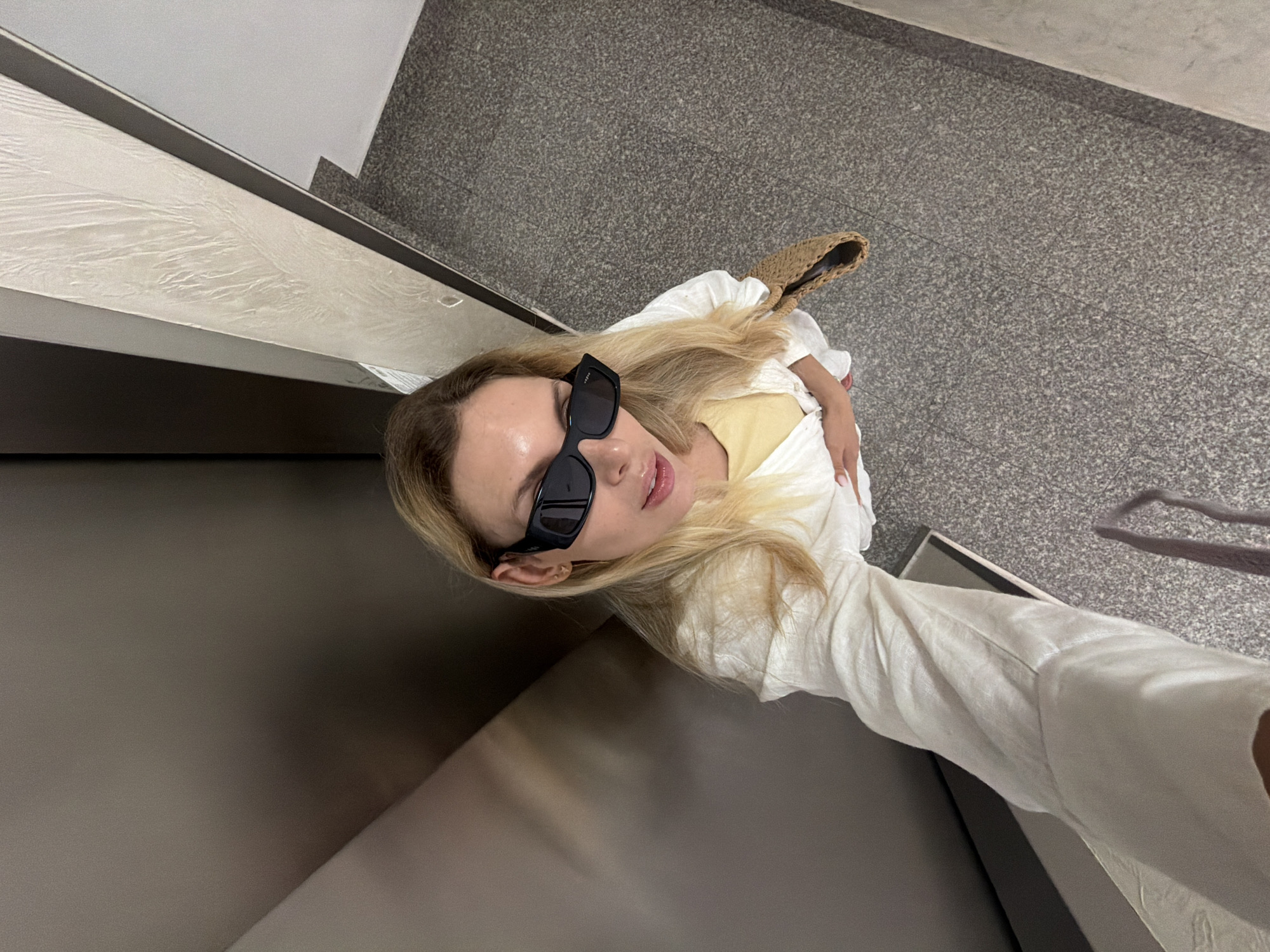 A woman is taking a selfie in an elevator in Budva, Montenegro. She is wearing black sunglasses, a white linen shirt over a yellow top, and carrying a straw bag. The elevator walls are a dark metallic color and a textured light beige. The floor is gray speckled granite. The lighting is bright and overhead.