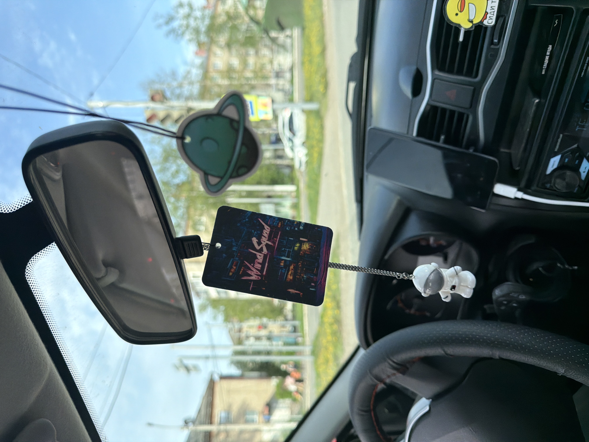 The image depicts the interior of a car, likely in the city of Chaikovsky, Russia, as suggested by the Cyrillic text on an air freshener. The view is from the driver's perspective, focusing on the dashboard, steering wheel, and rearview mirror.
A smartphone is mounted on the dashboard, charging with a white cable. A yellow duck air freshener hangs near the central vents. A "Wind Squad" air freshener and a small astronaut figurine are suspended from the rearview mirror.
Outside the car window, buildings, vegetation, and traffic lights are visible, hinting at an urban environment. A vehicle can be seen in the distance. The sky is partially visible, suggesting a sunny or partly cloudy day. The steering wheel is black with red stitching.