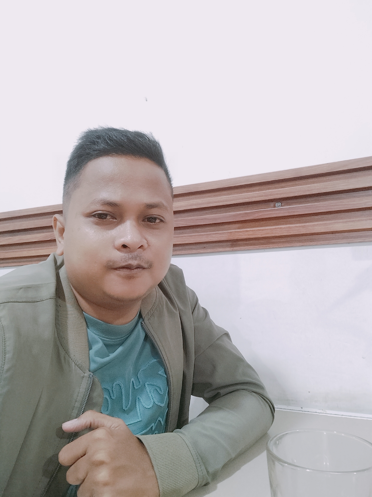 A medium shot selfie shows a man with short, dark hair and light brown skin seated at a table. He is wearing a teal t-shirt with a graphic design and a light olive green bomber jacket. His right hand rests on the jacket, near the zipper. A clear glass is visible on the table in front of him. The background consists of a white wall with a horizontal wooden paneling design. The lighting is soft, suggesting an indoor setting. The scene appears to be in Lhokseumawe, Indonesia, though specific context for the location is not provided in the image.
