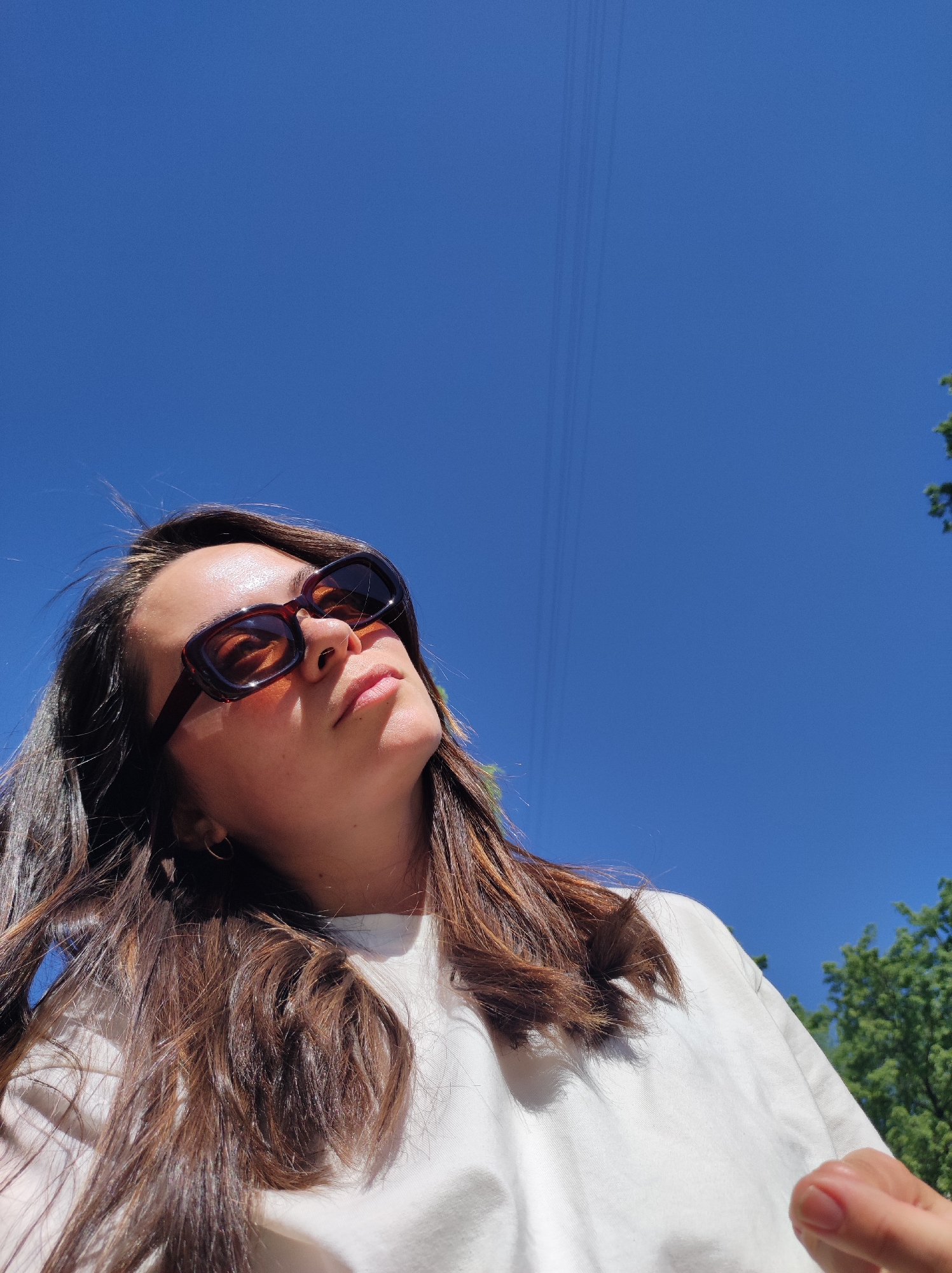 The image depicts a woman wearing sunglasses, looking upwards towards a bright blue sky. Some utility lines are visible against the sky. Trees appear in the periphery of the frame. The woman is wearing a white t-shirt. The exact location in the city of Chaikovsky, Russia, is not discernable from the image.