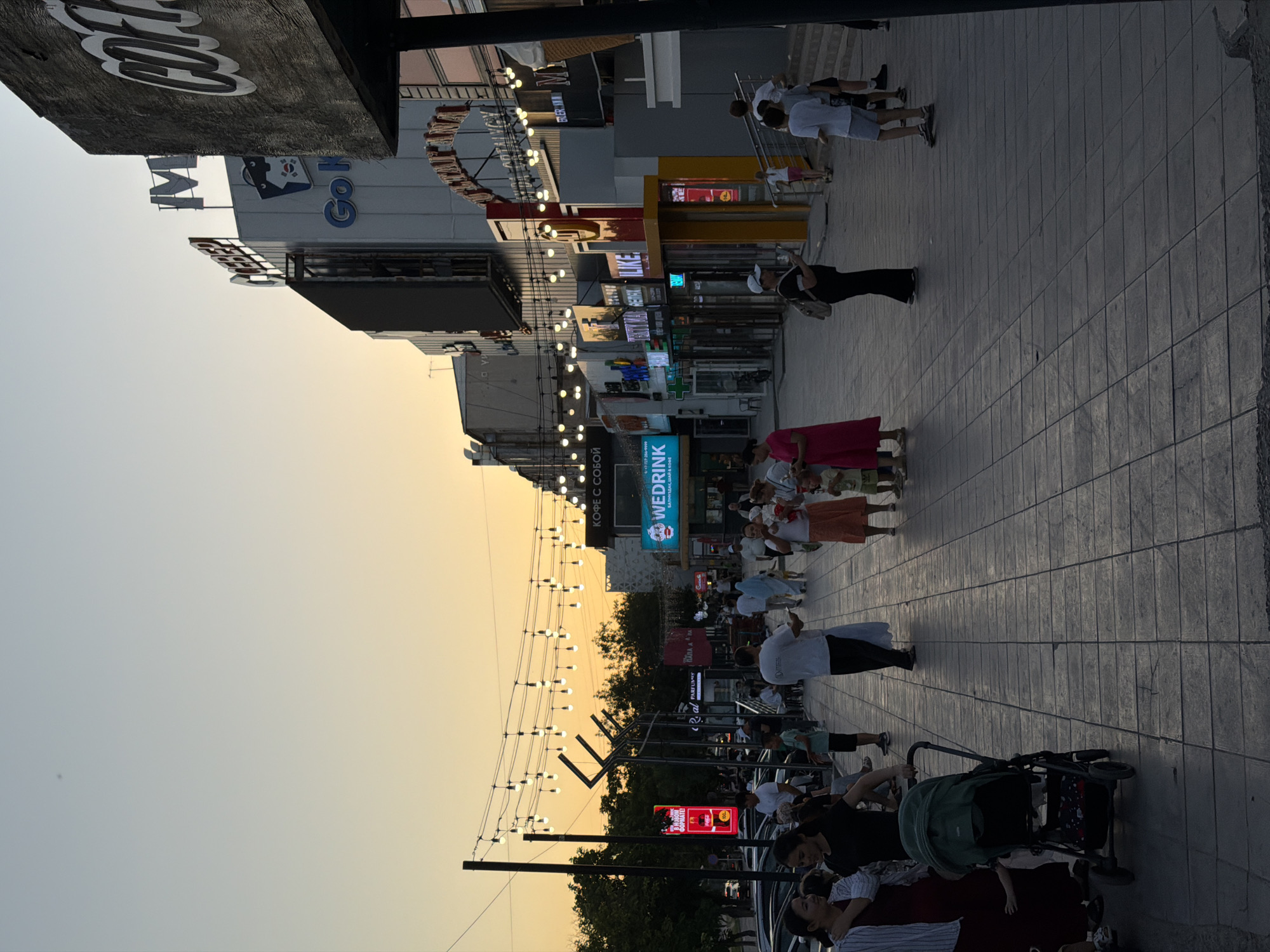 The image depicts a street scene in the city of Shymkent, Kazakhstan, during the evening. The sky is a pale orange and blue gradient, suggesting sunset or sunrise. The street is paved with large square tiles. On the right side of the street are multi-story buildings with illuminated signs, indicating shops and businesses. One sign prominently displays the word "COFFEE". On the left side of the street, trees are visible behind a row of parked cars.
A string of decorative lights with exposed bulbs stretches across the street, suspended between poles. Several people are walking along the sidewalk. In the foreground on the left, a woman in a dark dress and a woman in a red dress are standing near a green baby stroller. Further down the sidewalk, a group of people are gathered. On the right side of the image, several children are walking or standing. The overall atmosphere appears to be a public space with moderate pedestrian traffic.