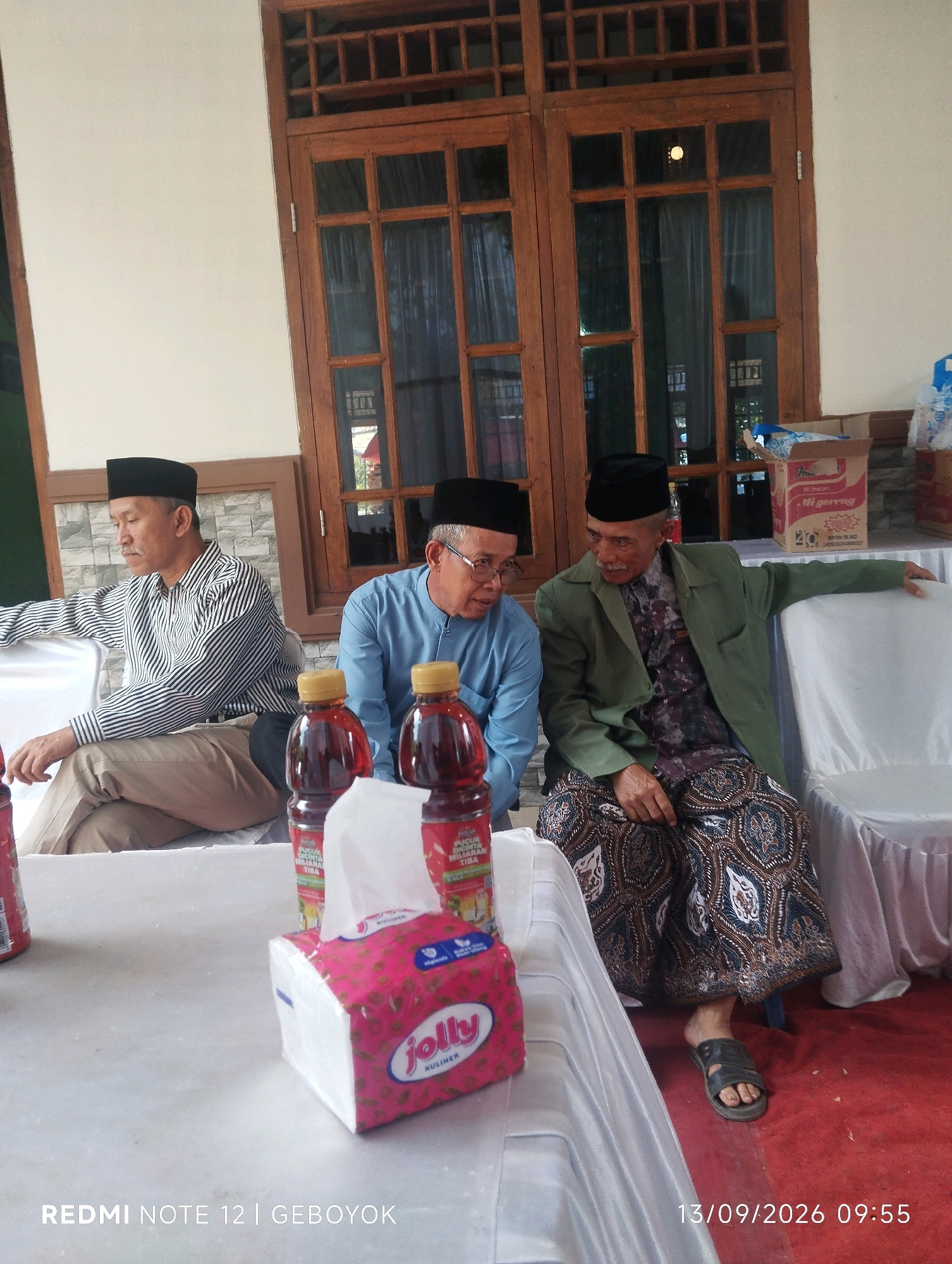Three men are seated on chairs in an outdoor setting, likely in Kuwukan, Indonesia. The man on the left wears a black peci, a striped long-sleeved shirt, and khaki pants. He is seated and looking to his left. The middle man, also wearing a black peci, has on a blue long-sleeved shirt and is looking down. In front of them on a table are two bottles of red liquid and a box of "Jolly" brand tissues. The man on the right is wearing a black peci, a green jacket over a patterned shirt, and a batik sarong. He is leaning back and looking to his right. The background features a wooden window with multiple panes and what appears to be a white wall. A cardboard box labeled "Mie Goreng" is visible to the right of the window. The seating area is set up with white chairs and a red carpet on the floor. The timestamp "13/09/2026 09:55" and "REDMI NOTE 12 | GEBOYOK" are watermarked in the lower right and left corners respectively.