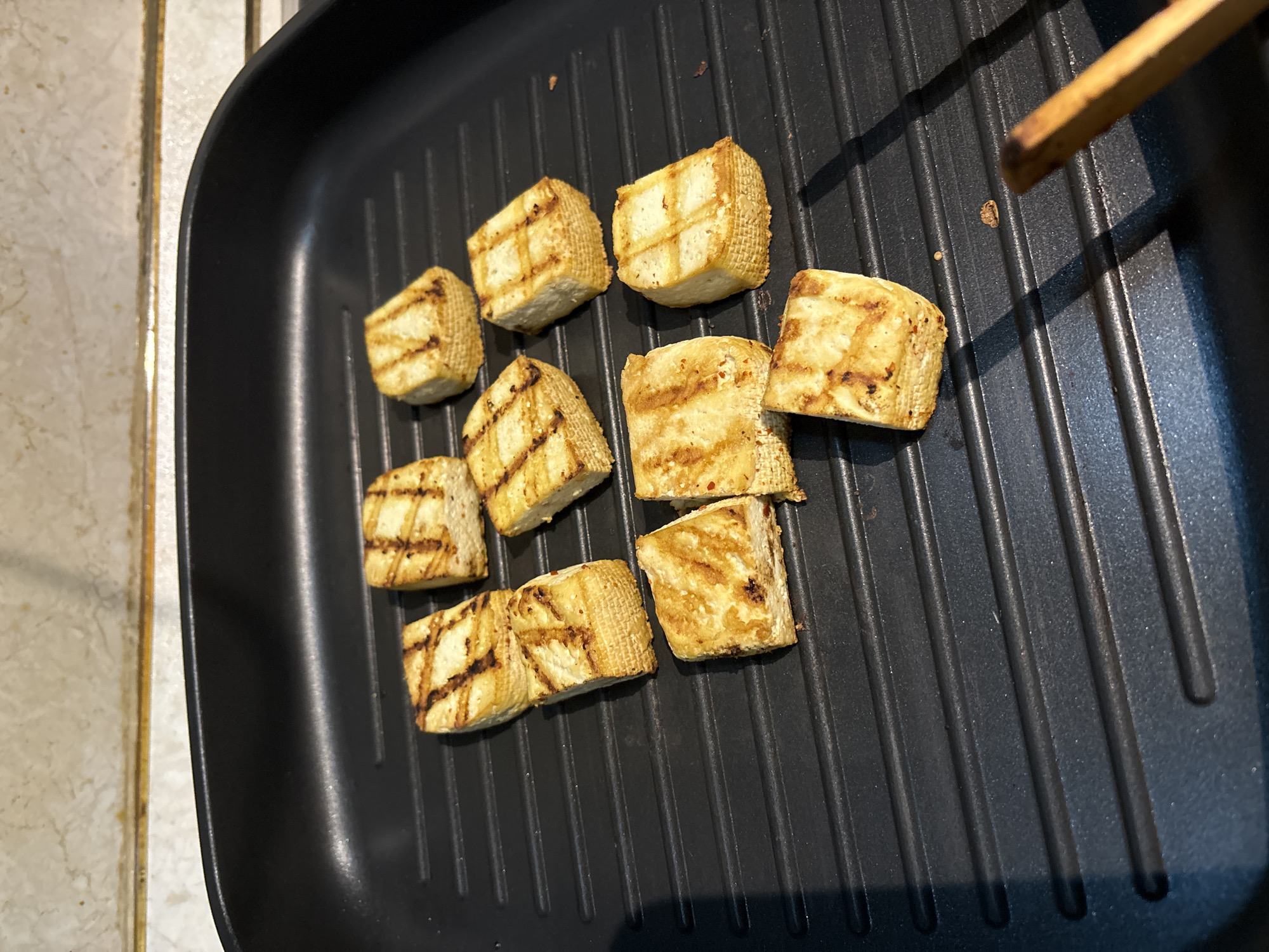 A black grill pan sits on a tiled surface. Inside the pan are approximately 10 tofu cubes, arranged somewhat randomly. The tofu cubes are cooked and have grill marks on their surfaces. A brown wooden utensil is partially visible in the upper right corner of the frame, presumably used for cooking or manipulating the tofu. The location appears to be a kitchen, as evidenced by the tiled surface and the presence of a cooking utensil and pan. The city of Unknown, Unknown is not discernible from the provided information.