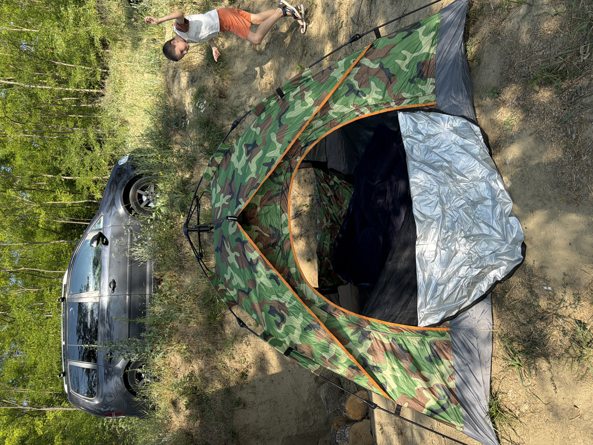 In Găleștii Noi, Moldova, a camouflaged tent is set up on sandy ground, partially covered by a silver tarp. Behind the tent, a dark gray station wagon is parked on a dirt incline, with its roof rack visible. A young boy, wearing a white t-shirt and orange shorts, is seen running and gesturing in the background, near the car and the incline. The environment is wooded, with a dense canopy of green trees and dry vegetation.