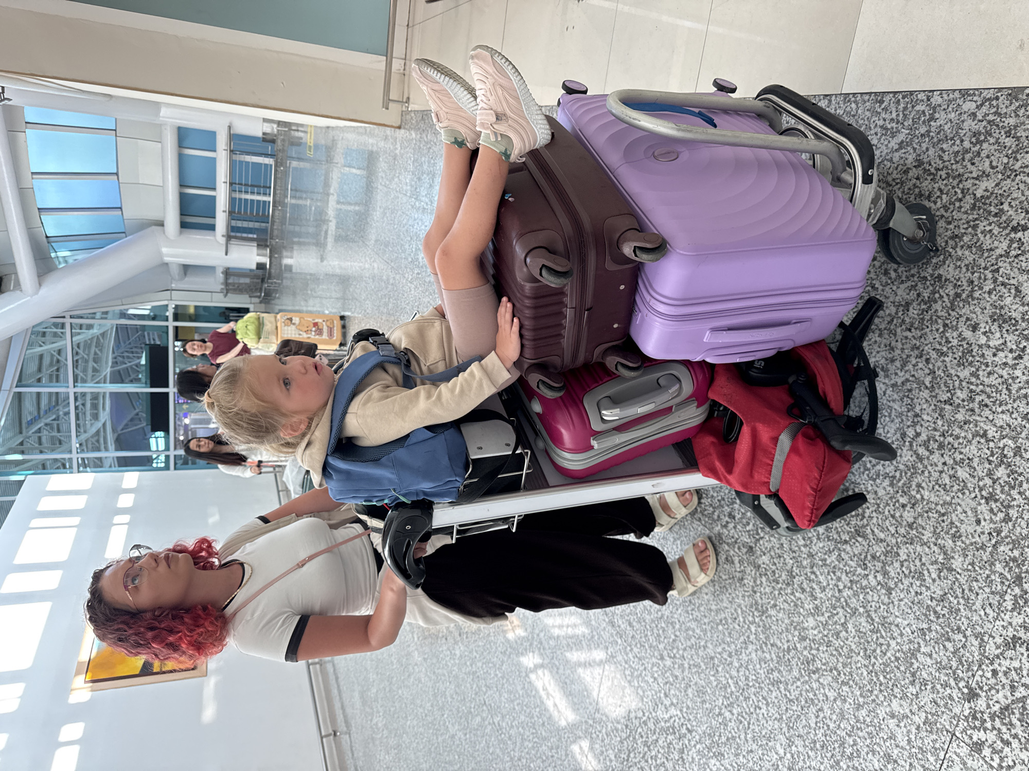 The image captures an indoor scene within a modern airport terminal in Kuta, Indonesia. In the foreground, a woman and a young child are positioned next to a heavily loaded luggage cart.
The woman, located on the left, appears to be in her late 20s or early 30s, with shoulder-length, curly hair dyed red and pink. She wears gold-rimmed cat-eye glasses and is dressed in a white t-shirt with black trim, black baggy pants, and light beige two-strap sandals. A light-colored garment is draped over her shoulders, and a thin gold chain necklace is visible. Her left hand rests on the black handle of the luggage cart, which she is pushing. Her gaze is directed upwards and to the right.
To the right of the woman, a young child, estimated to be between 2 and 4 years old, sits on the stacked luggage. The child has blond hair, visible blue eyes, and wears a beige hooded sweatshirt, beige shorts, and pink sneakers with white soles and velcro straps, along with light-colored, patterned socks. A small blue backpack is worn on the child's back. The child's legs are crossed, and hands rest on the luggage.
The silver-framed luggage cart carries multiple suitcases. The child is seated on a dark purple hard-shell suitcase, which is stacked above a light purple hard-shell suitcase. A pink hard-shell suitcase is partially visible on the far right side of the cart, beneath the dark purple one. All visible suitcases feature spinner wheels. A red soft-sided bag is secured to the lower front section of the cart's frame.
The environment features a speckled grey terrazzo floor in the foreground, transitioning to a lighter beige tile section on the far right. The background shows tall white walls, large white structural pillars, and expansive windows with blue-tinted panels. An abstract rectangular artwork with orange and brown hues is mounted on the white wall in the upper left background. In the mid-ground, several other individuals are present, including a woman behind the child's head, carrying a light green bag next to a small yellow wheeled suitcase with a teddy bear design. Metal railings are also visible in the mid-ground. The area is brightly lit.
