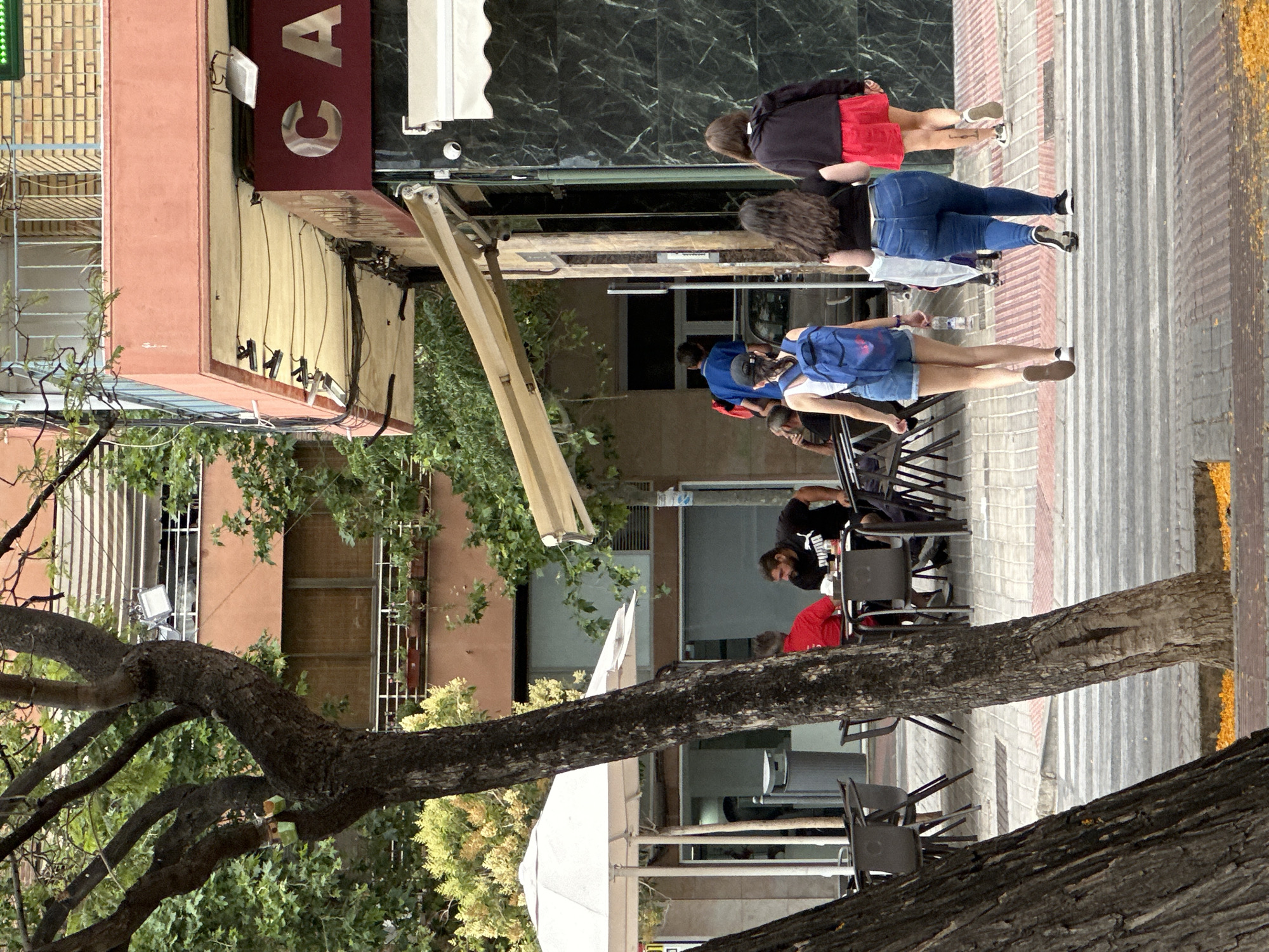 In an urban environment, likely in the city of Unknown, Unknown, a street scene unfolds with several individuals and a cafe. The perspective is from ground level, looking towards a street and sidewalk.
In the foreground, the trunk of a large tree with dark, textured bark dominates the left side of the frame. Its branches extend upwards and outwards, partially obscuring the background. To the right, a crosswalk with white stripes is visible on the street. Scattered yellow petals lie on the sidewalk and the edge of the street.
On the sidewalk, a cafe with outdoor seating is apparent. White umbrellas cover tables and chairs, some of which are occupied. Two men are seated at a table on the right side of the cafe area. One man wears a red shirt and the other a black t-shirt with a white logo. They appear to be engaged in conversation.
Further down the sidewalk, closer to the cafe's entrance, a group of people are walking away from the camera. Three women are prominent. The woman in the front is wearing denim shorts and a white tank top, carrying a blue backpack. The woman in the middle is in blue jeans and a black top. The woman on the far right wears a red skirt and a black top. Behind them, a man in a blue shirt is walking, and further back, another person is partially visible. The cafe itself has a dark, reflective facade with a large red sign displaying the letters "CA" in silver. A white awning extends over the entrance.
The background features buildings with multiple stories, some with balconies and windows, interspersed with trees and foliage. The overall impression is a typical street scene with people going about their day near a cafe.