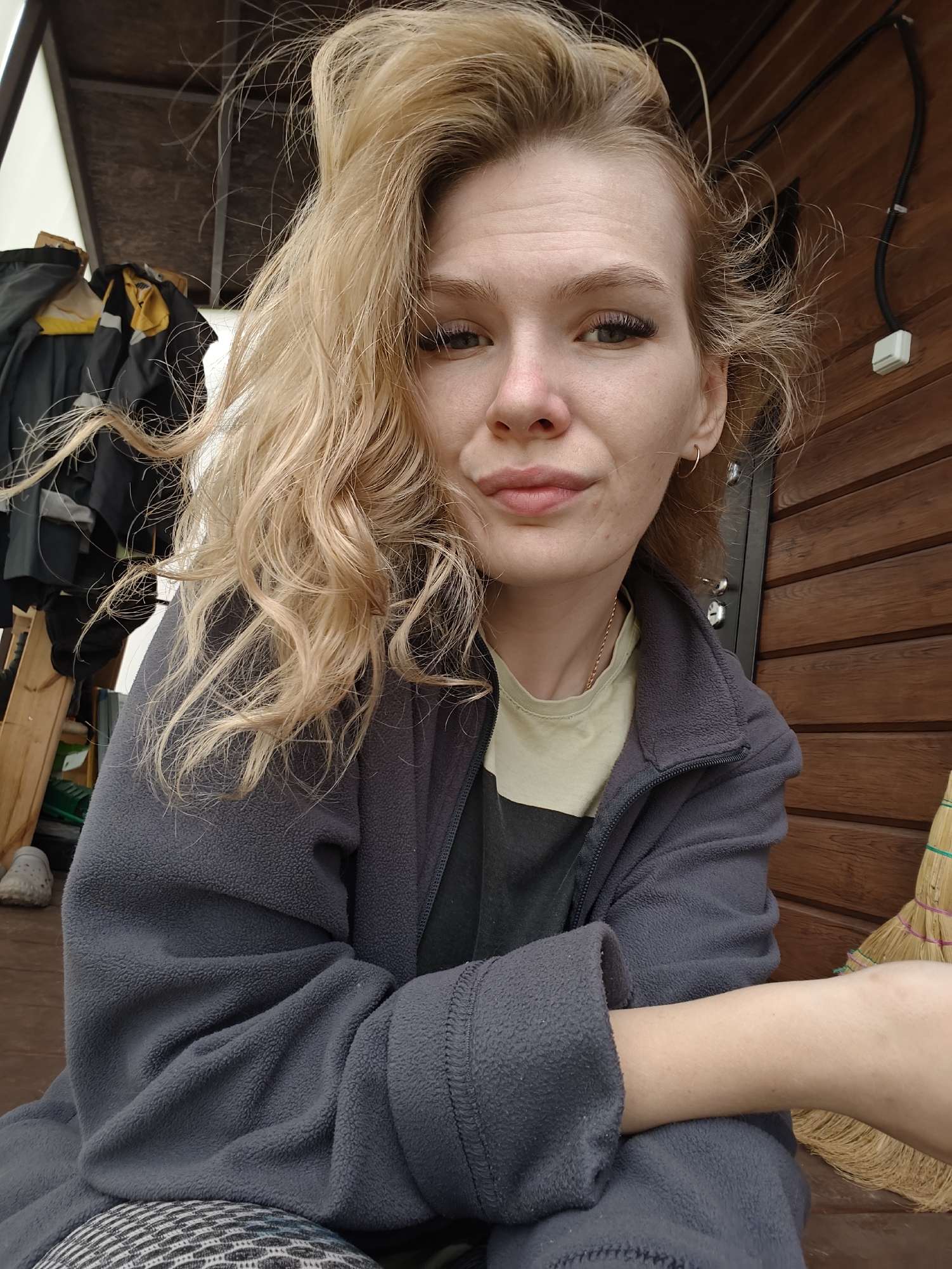 A young woman with blonde, wavy hair is looking at the camera, smiling slightly. She is wearing a grey fleece jacket over a light-colored t-shirt. Her arms are crossed in front of her. The background shows a wooden wall and some hanging clothing. The scene appears to be outdoors, possibly on a porch or in a sheltered area. There is a broom leaning against the wooden wall. The location is identified as Русский Вожой, Russia.