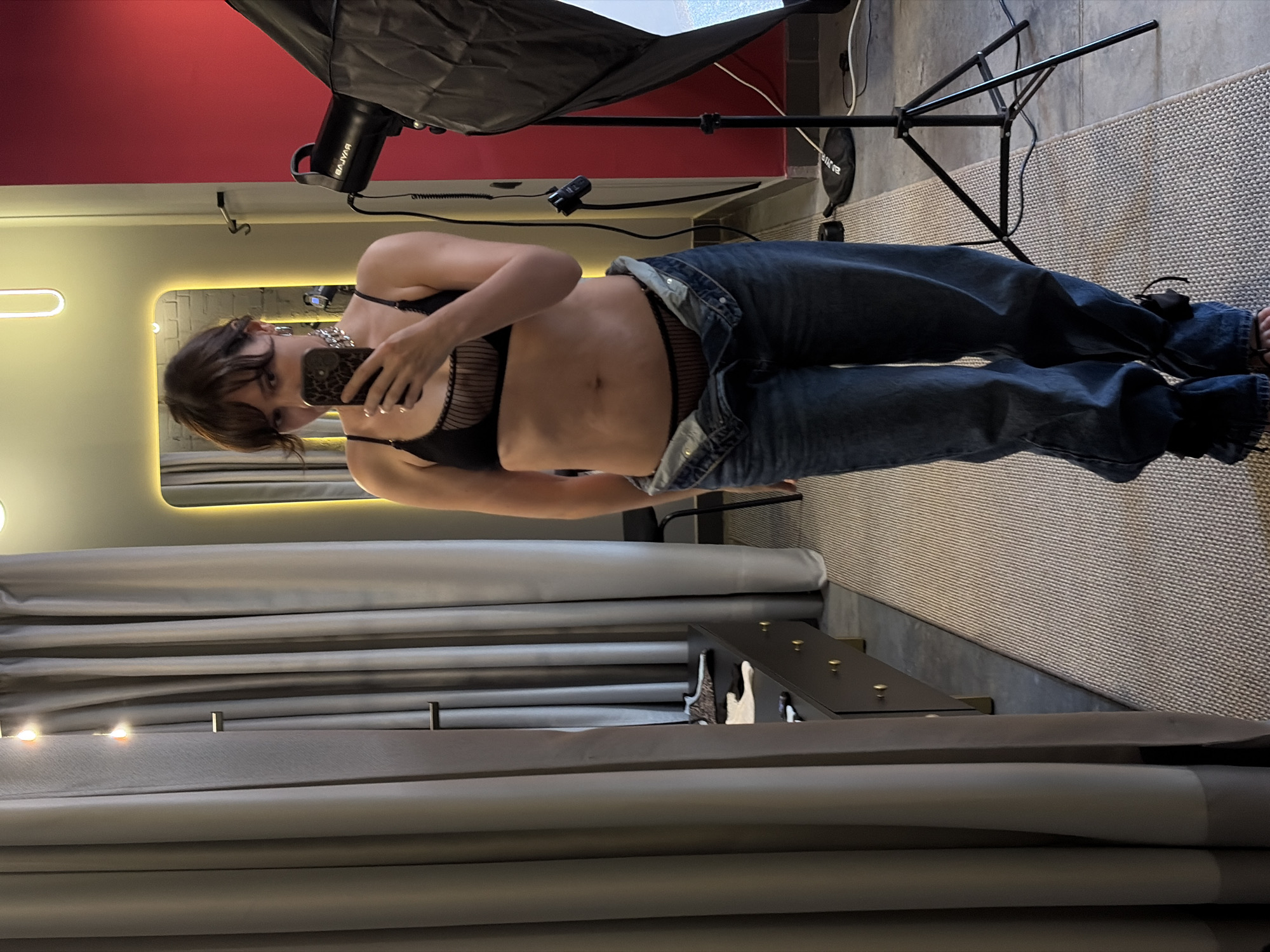 In an indoor studio setting, a woman with dark hair and bangs is taking a mirror selfie. She is wearing a black bralette with thin straps and a sheer, striped mesh detail. Her jeans are unbuttoned and slung low on her hips, revealing matching sheer mesh underwear. She is also wearing a chunky silver choker necklace and hoop earrings. The room features light-colored curtains to the left, a dresser with gold knobs, and a large, brightly lit mirror reflecting the scene. To the right, a red wall is visible behind a professional photography light stand with a softbox. The floor is covered with a patterned rug. The woman is holding a smartphone with a leopard print case. There are no obvious signs indicating the location is in Chaikovsky, Russia.