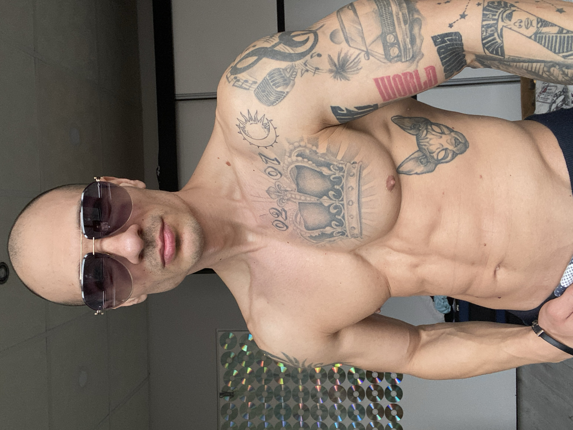 A bald, muscular man with numerous tattoos on his chest and arms is posing indoors. He is wearing sunglasses. The location is not identifiable as Pleven, Bulgaria, based on the image content.