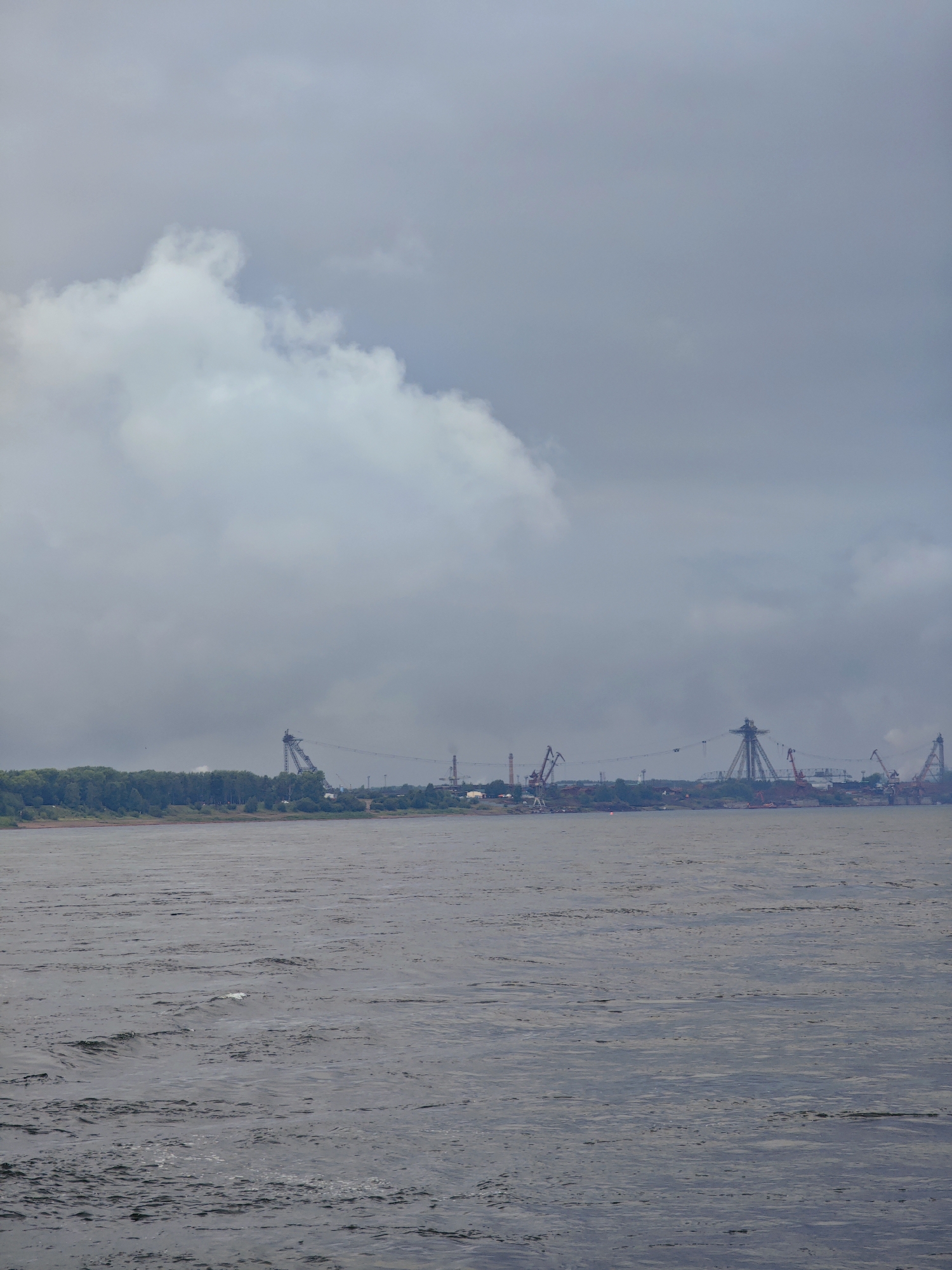 A wide, dark gray river dominates the foreground of the image, with visible ripples and small waves suggesting movement. In the middle ground, a dense, dark green treeline stretches across the horizon, forming the bank of the river. Beyond the trees, a hazy industrial landscape is visible, characterized by numerous tall cranes and industrial structures. These structures are mostly in shades of red and gray, with a large, skeletal tower-like structure prominently featured. The sky above is overcast with heavy, light gray clouds, obscuring any direct sunlight and contributing to the overall muted and somewhat somber atmosphere of the scene. There are no visible people in the image. This scene is consistent with the industrial port areas along the Yenisei River in Lesosibirsk, Russia, known for its timber industry and related infrastructure.