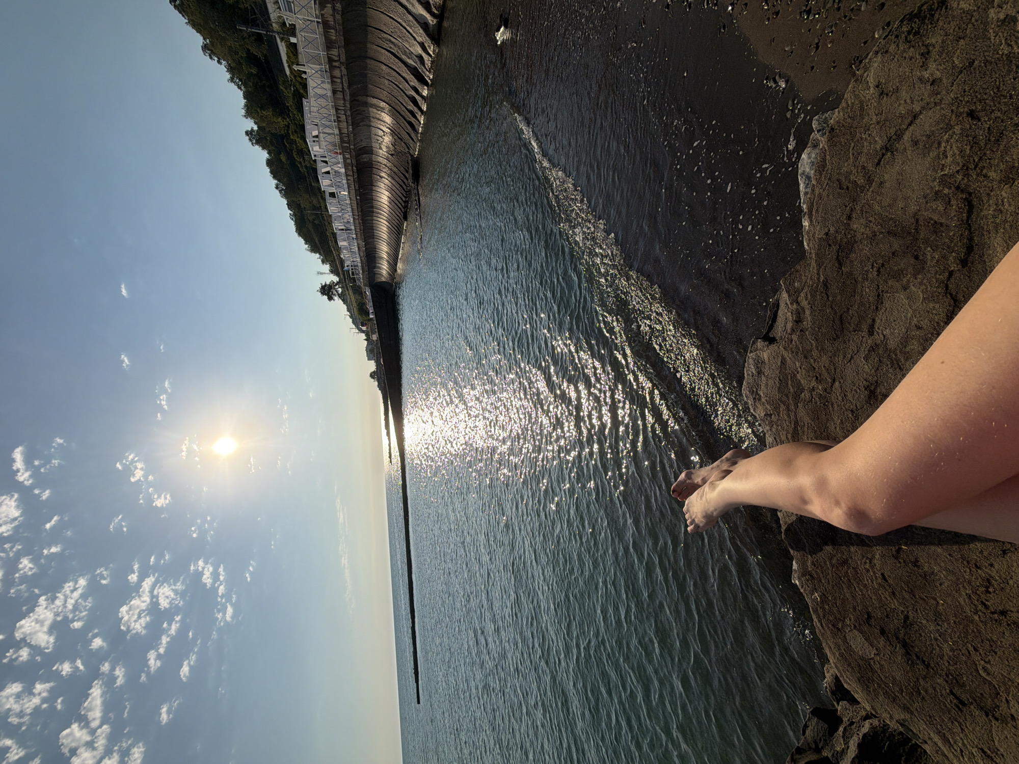 The image shows a view of the Black Sea coast in Sochi, Russia, on a sunny day. The sun is shining brightly, casting a shimmering reflection on the water. In the foreground, a person's legs are visible, resting on a rock. The legs are tanned and appear to be bare. The person is facing away from the camera, towards the sea.
The background features a curved seawall with a white fence and white tents, suggesting a beach resort or a public beach area. Lush green trees line the hillside behind the seawall. The water is a deep blue, with gentle waves lapping at the dark-colored shore. A long, narrow breakwater extends into the sea in the distance. The sky is clear with scattered white clouds. The overall impression is one of relaxation and enjoyment of a seaside environment.