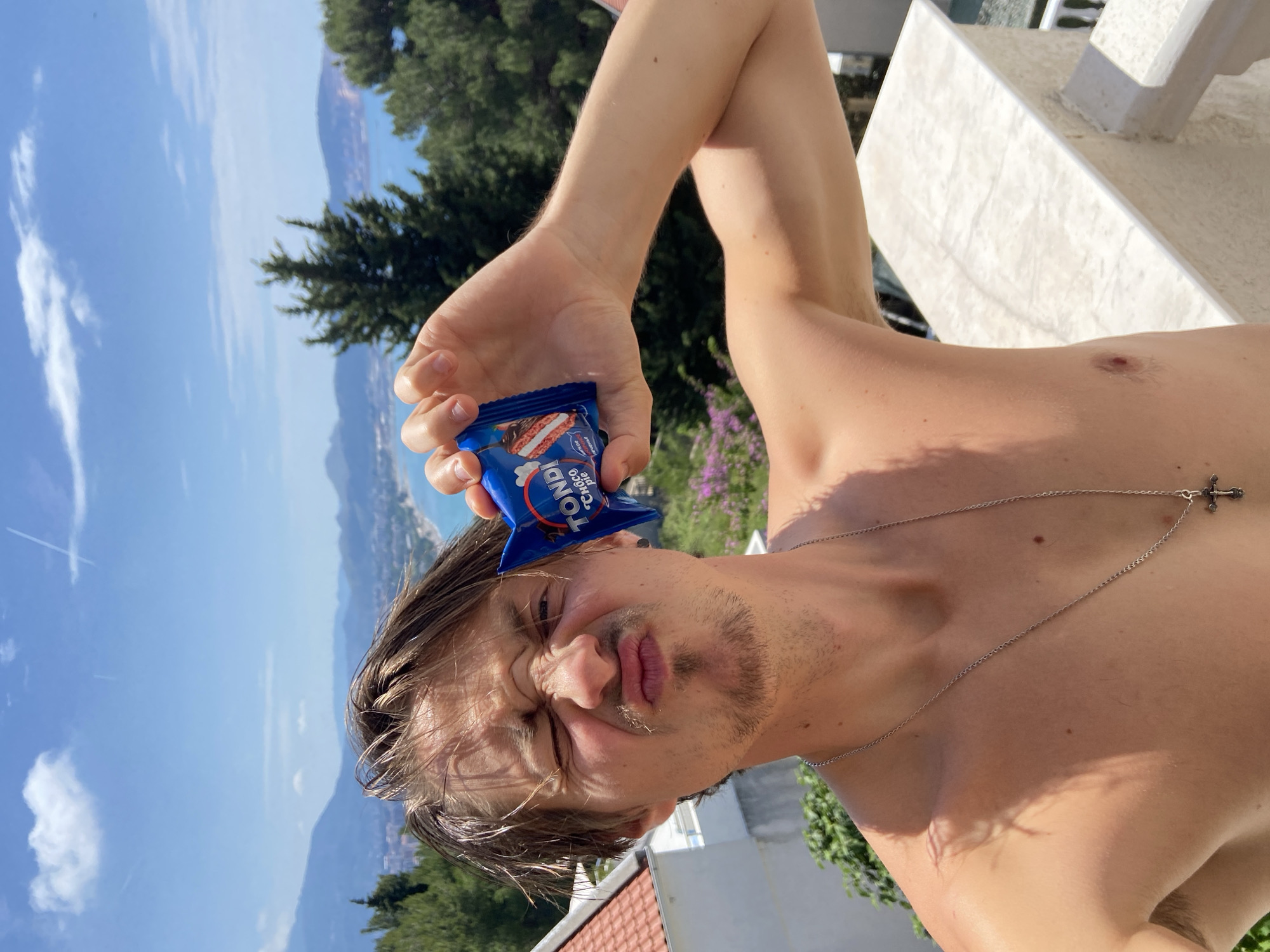 A shirtless man with light brown hair, a light beard, and a silver chain necklace with a cross pendant is holding a small, blue package of "TONDI Choco Pie" in front of his right eye. He is squinting and making a funny face. The background consists of a landscape with trees, a body of water, and a blue sky with clouds. The location appears to be Šušanj, Montenegro, based on the typical landscape.