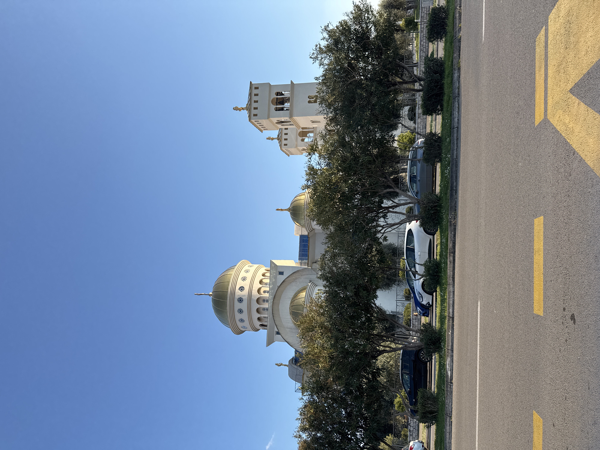 The image captures a view of the St. Jovan Vladimir Orthodox Cathedral in Bar, Montenegro. The cathedral, characterized by its white facade, golden domes, and a bell tower, stands adjacent to a road marked with yellow lane markings. Several trees partially obscure the building. Parked cars are visible along the roadside. The sky is clear and blue.