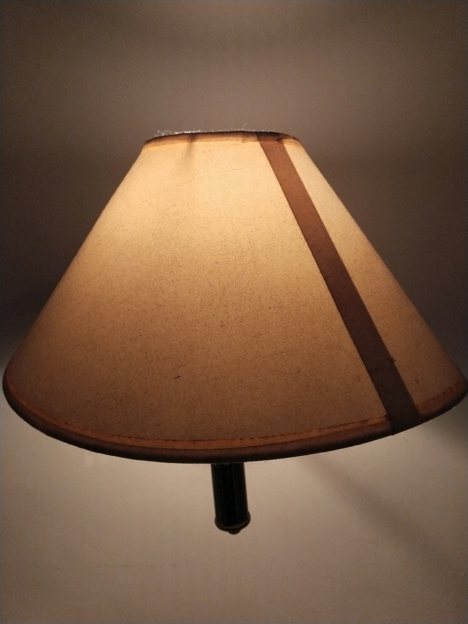 A beige lampshade with a darker vertical stripe is illuminated from within. The lampshade is conical in shape and hangs from a black lamp base, visible at the bottom of the image. The background is a gradient of dark to light, with the top of the image being a darker brown and the bottom a lighter beige. The scene is indoors and likely a domestic setting. There is no indication of the location being in Sinduadi, Indonesia.