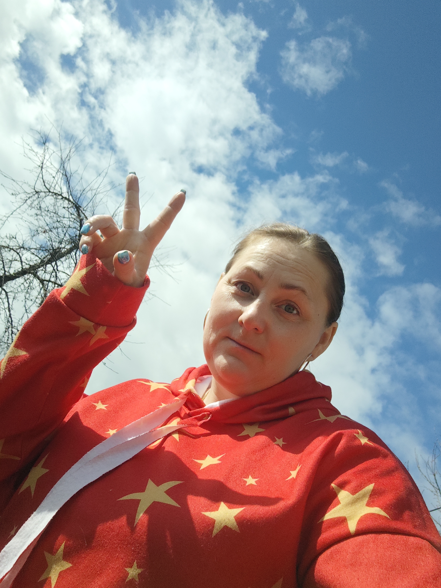 A low-angle shot depicts a woman in a red hoodie with gold stars, holding up a "peace" sign with her left hand. She appears to be outdoors, based on the blue sky and scattered white clouds in the background. Bare tree branches are visible in the upper left corner. The photo was taken in the city of Фоки, Russia.