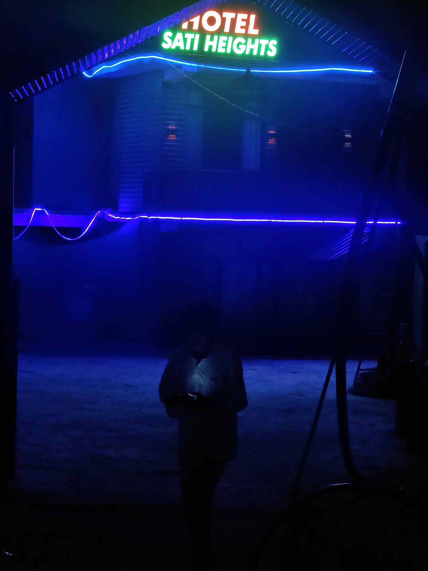 The image captures the exterior of "HOTEL SATI HEIGHTS" in Barheta, India, at night. The hotel's name is illuminated by neon signs, with "HOTEL" in orange and "SATI HEIGHTS" in green, positioned above an entranceway framed by blue neon lighting. The building appears to be constructed of brick and is dimly lit, with the blue lights casting a strong glow on the surrounding area. In the foreground, a person is silhouetted against the light, holding a mobile phone. The ground appears to be a paved or dirt surface, and the overall atmosphere is dark and somewhat mysterious due to the low light conditions and the vibrant neon signage.