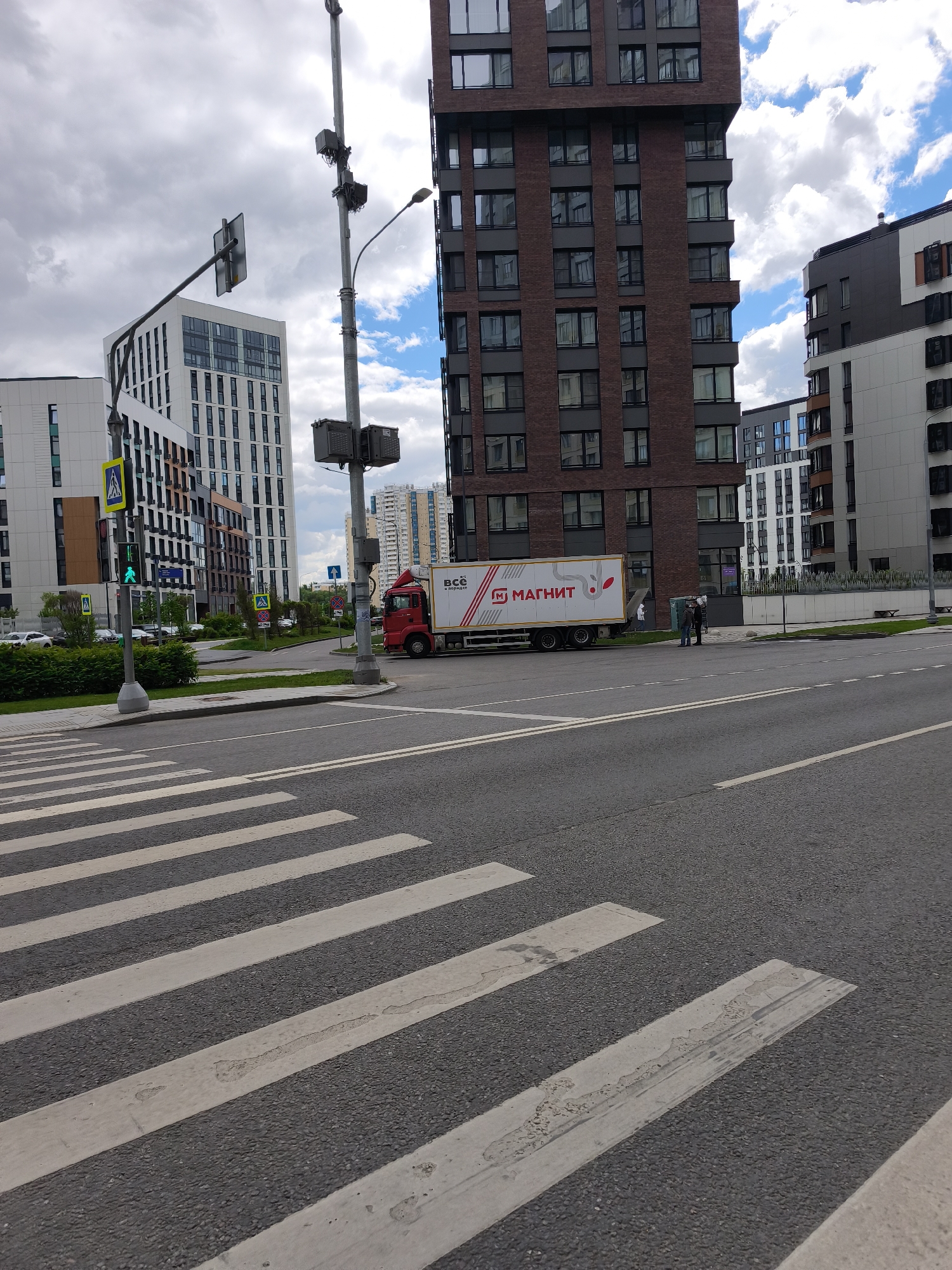 The image shows a street scene in Moscow, Russia, featuring a crosswalk in the foreground. A red delivery truck with "Магнит" (Magnit) branding is parked on the side of the road, possibly near a delivery point. Two people are standing near the back of the truck. Several modern apartment buildings are visible in the background with unique architectural designs. A pedestrian traffic light shows a green signal. Other street furniture, including utility poles and road signs, are present. The sky is overcast with scattered clouds.