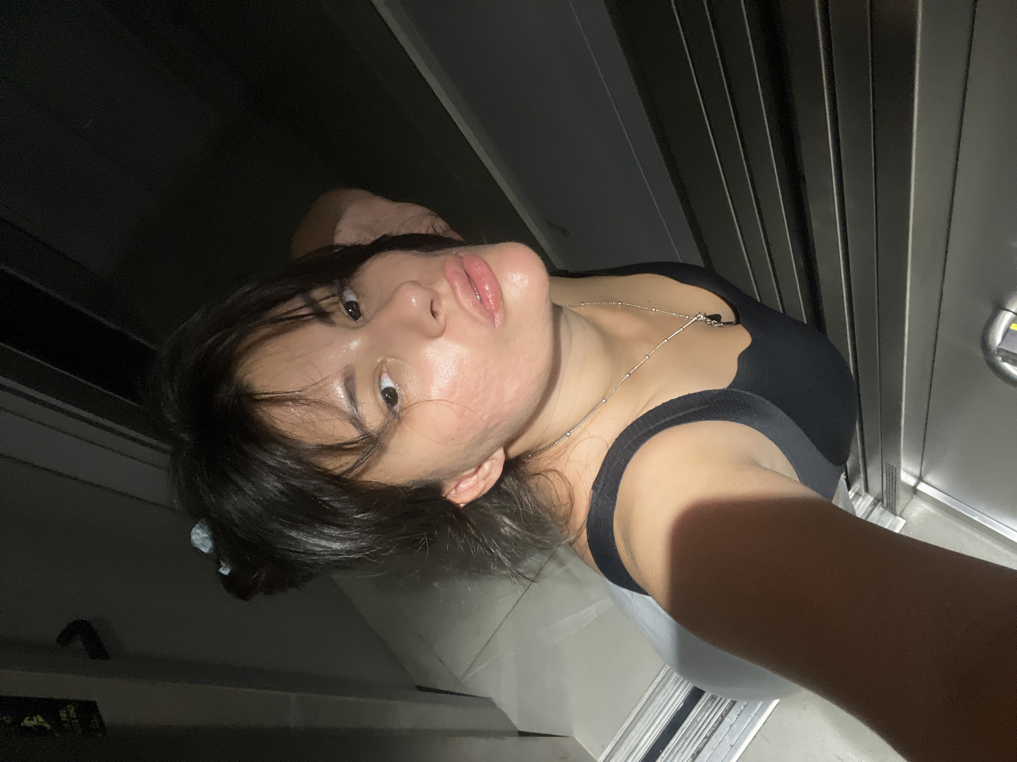 The image is a selfie taken in what appears to be an elevator or a similar enclosed space with ribbed metallic walls. A young woman with dark hair and a black tank top is the sole subject. She is looking directly at the camera with a neutral expression. A silver necklace with a small pendant is visible around her neck. The lighting is dim, suggesting it might be nighttime or an interior setting with limited light. The background is dominated by the vertical lines of the metallic walls, creating a sense of confinement. There are no visible signs of text or specific environmental cues that would indicate the exact time of day or weather conditions. The setting seems utilitarian and nondescript.