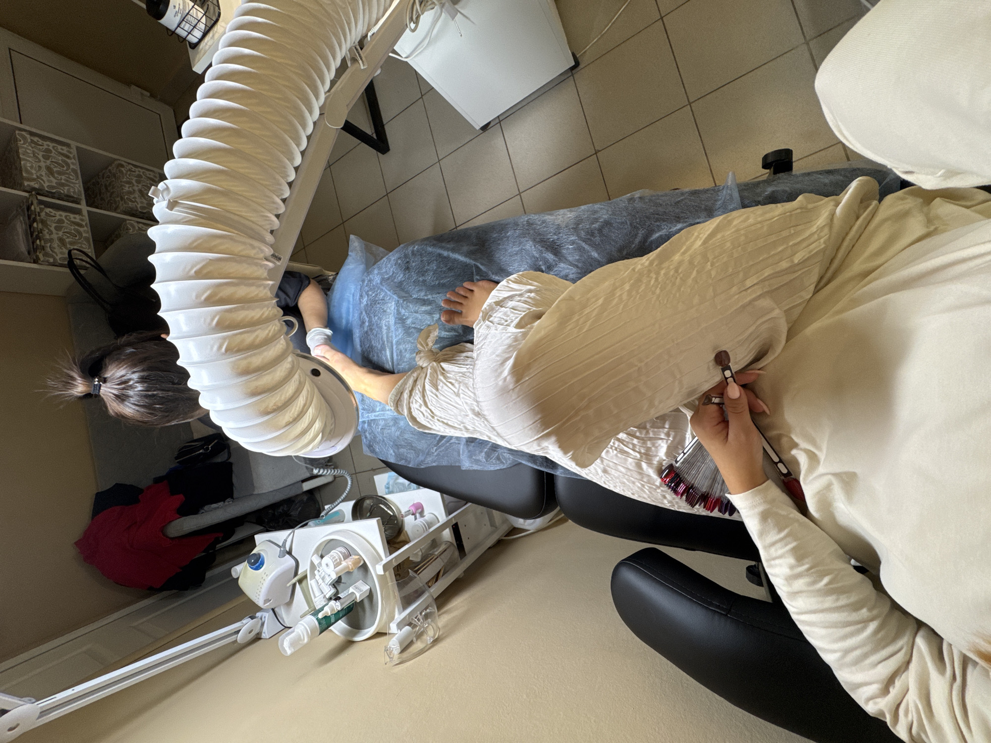 The image shows a person in a nail salon, likely in the city of Chaikovsky, Russia. The person is seated in a black chair, with their left leg extended and resting on a blue disposable mat. They are wearing a cream-colored long-sleeved shirt and matching pants. A nail technician, with their back to the camera, is working on the person's foot, wearing white gloves and a dark top. A large, white, flexible exhaust tube is positioned over the technician's workspace, presumably to remove dust and fumes. In the foreground, the person is holding a nail color swatch with various shades of red and purple. To the left, there is a white manicure station with various bottles and tools, including what appears to be a nail drill. The room has tiled flooring and a wall with a light-colored finish. In the background, there is shelving with decorative boxes, and a red garment is draped over a rack. The overall impression is of a clean and functional nail salon.