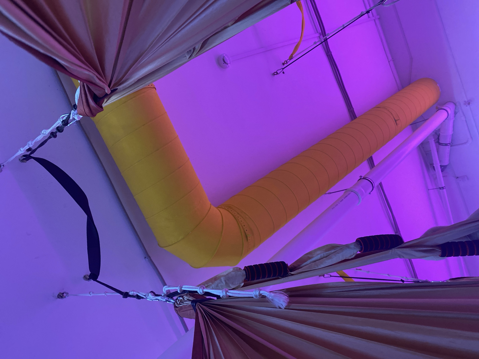 The image is taken from a low angle, looking up at the ceiling of a room in Chaikovsky, Russia. The ceiling is predominantly white and illuminated with a purple/blue light. Prominently featured are various pieces of aerial yoga equipment, including fabric hammocks in shades of tan/brown and a large, yellow, L-shaped ventilation duct that runs along the ceiling. The hammocks are suspended from the ceiling by white ropes and black straps attached to fixtures. Visible in the background are white pipes and electrical conduits also attached to the ceiling.