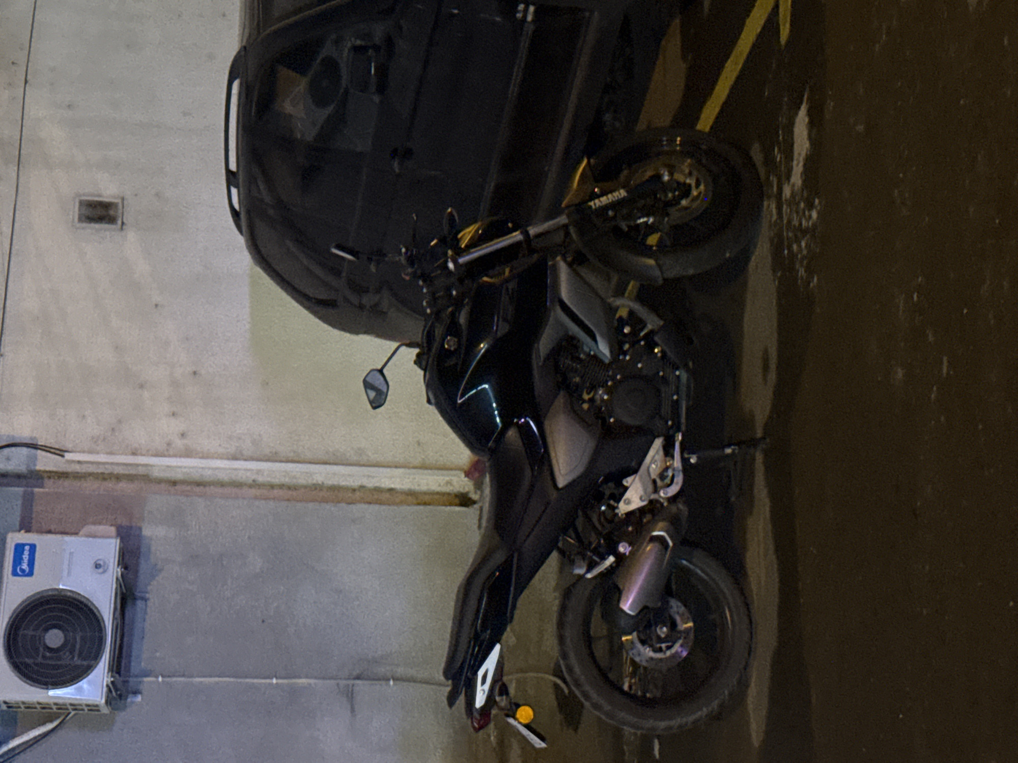 A black Yamaha motorcycle is parked next to a dark-colored SUV in a dimly lit outdoor area. The motorcycle is positioned at an angle, with its front wheel slightly turned. A silver air conditioning unit is mounted high on a gray concrete wall to the left of the motorcycle. The ground appears wet and reflective. The scene is set in an urban environment, possibly a parking lot or alleyway, in the city of Tbilisi, Georgia.