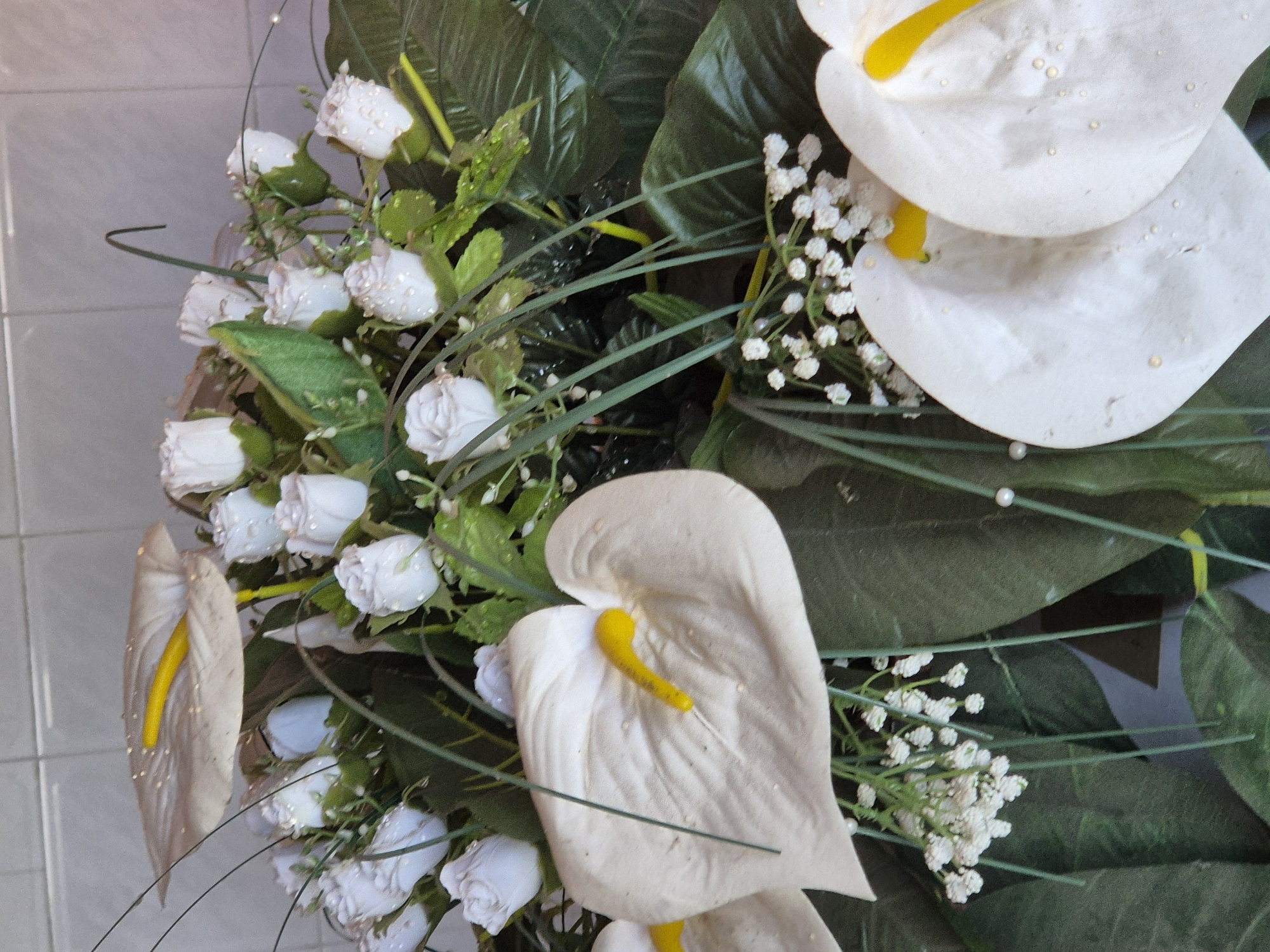 The image shows a floral arrangement, primarily composed of white roses and white anthurium lilies, along with smaller white filler flowers and green foliage. The roses and anthuriums appear to be artificial, possibly silk or plastic. The arrangement includes decorative pearl accents. The background suggests an indoor setting with tiled walls. The image's location in Pavlikeni, Bulgaria, is not directly discernable from the visual information.
